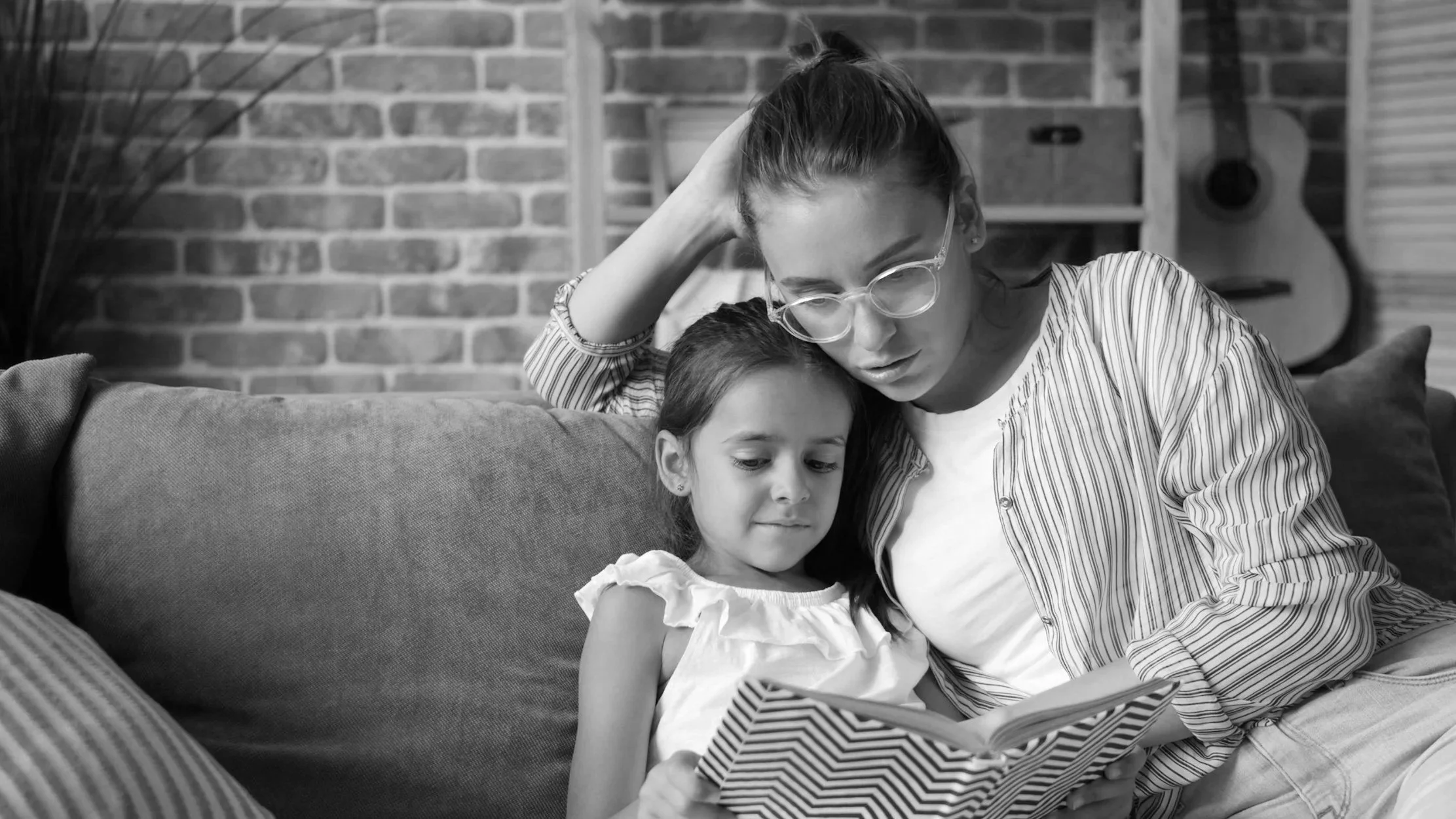 A woman and a young girl sitting on a couch, reading a book together. The woman has her arm around the girl and appears focused on the book. The background includes a brick wall and a guitar.