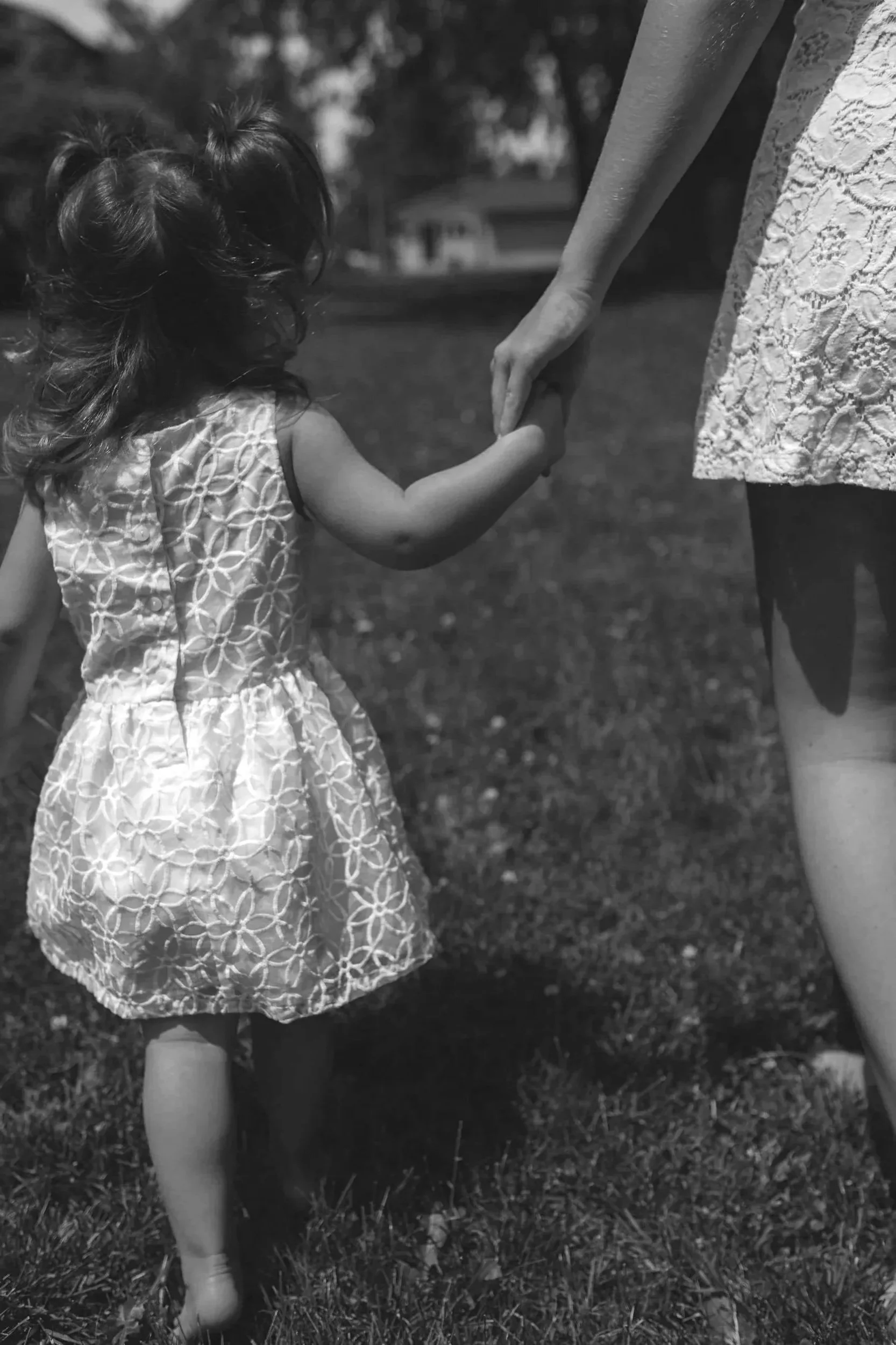 A young girl with curly hair wearing a floral dress, holding hands with an adult, walking on grass outdoors.