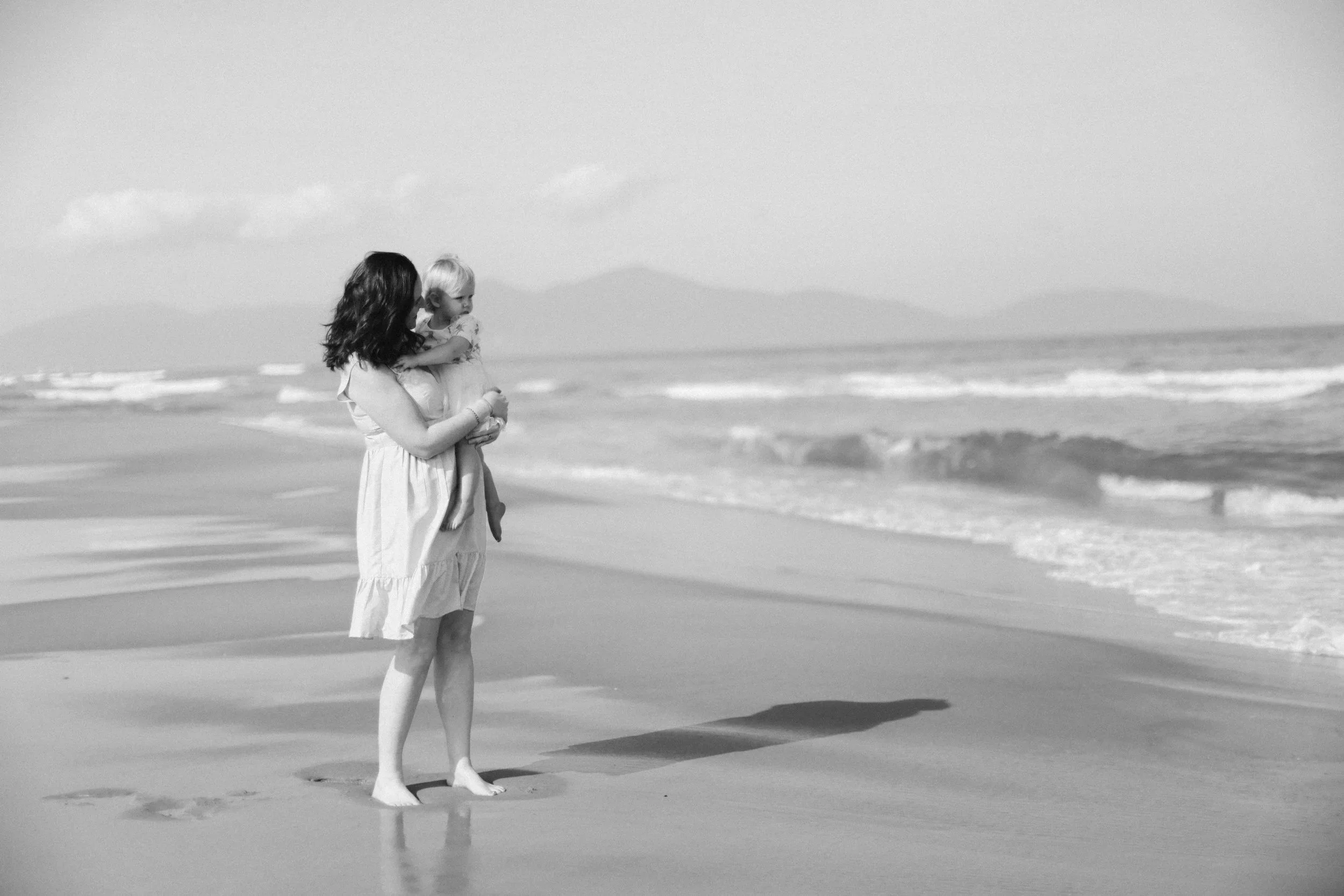 A woman holding a young child on a beach, with ocean waves and distant mountains in the background, in a black and white photo.