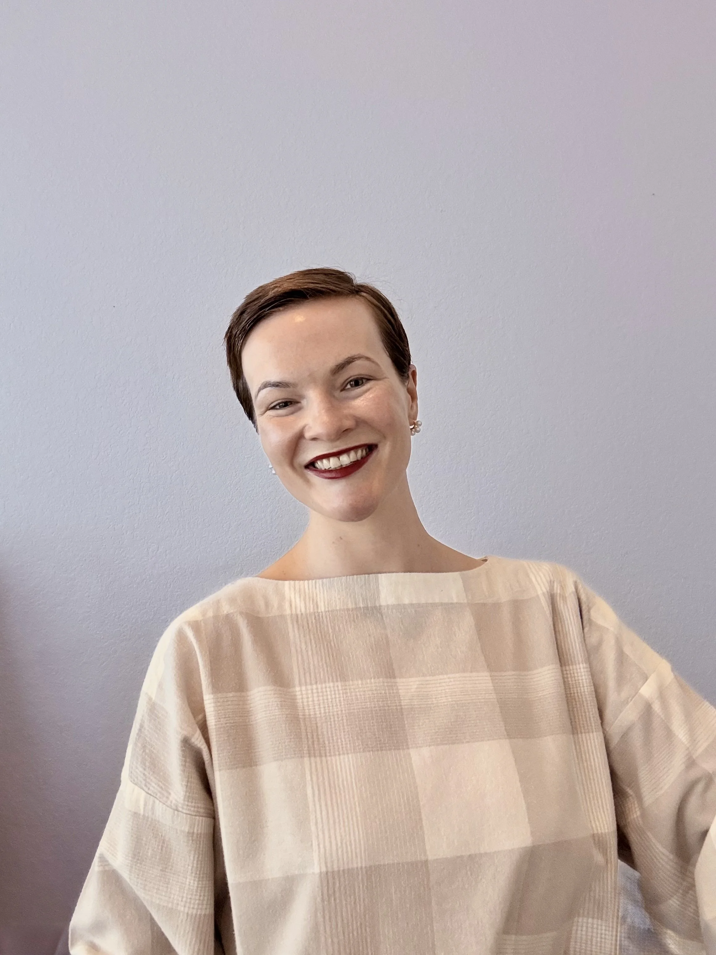 A woman with short brown hair smiling, wearing a beige checkered top, standing against a plain light gray wall.