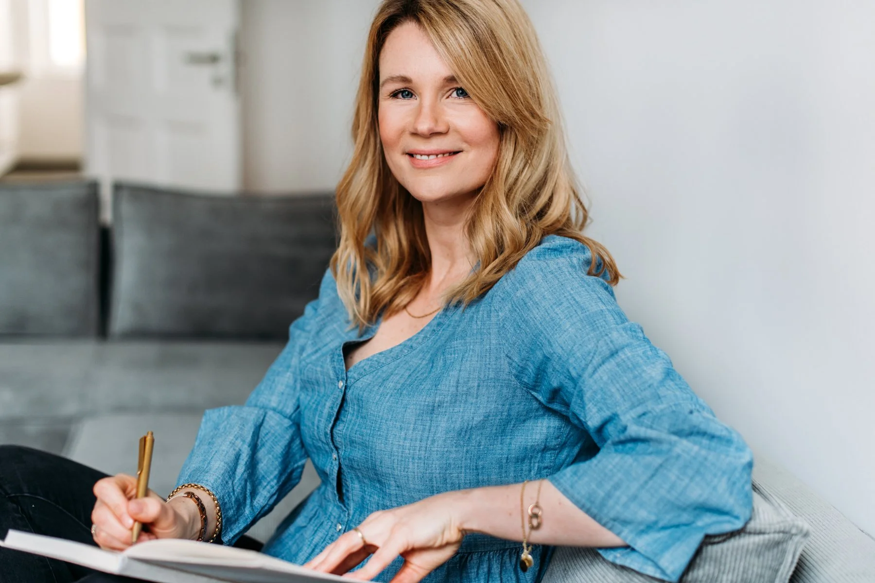 Junge Frau mit blonden, gewellten Haaren, trägt eine blaue Bluse, sitzt auf einem Sofa, schreibt in ein Notizbuch, lächelt und schaut begeistert.