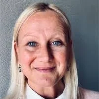 Nahaufnahme einer Frau mit blondem Haar und blauen Augen, lächelnd, vor einem grauen Hintergrund.