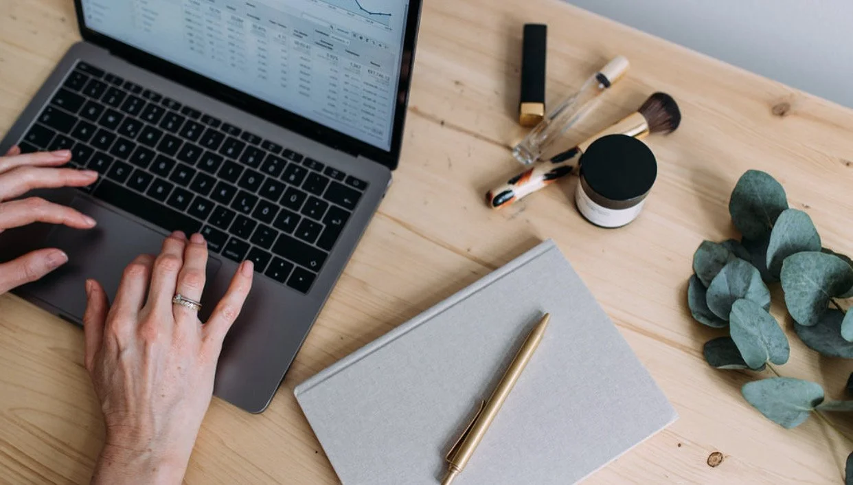 A person typing on a laptop keyboard on a wooden desk, with makeup brushes, a small jar container, a tube of lipstick, a glass perfume bottle, a plant, a gray notebook, and a gold pen on the desk.