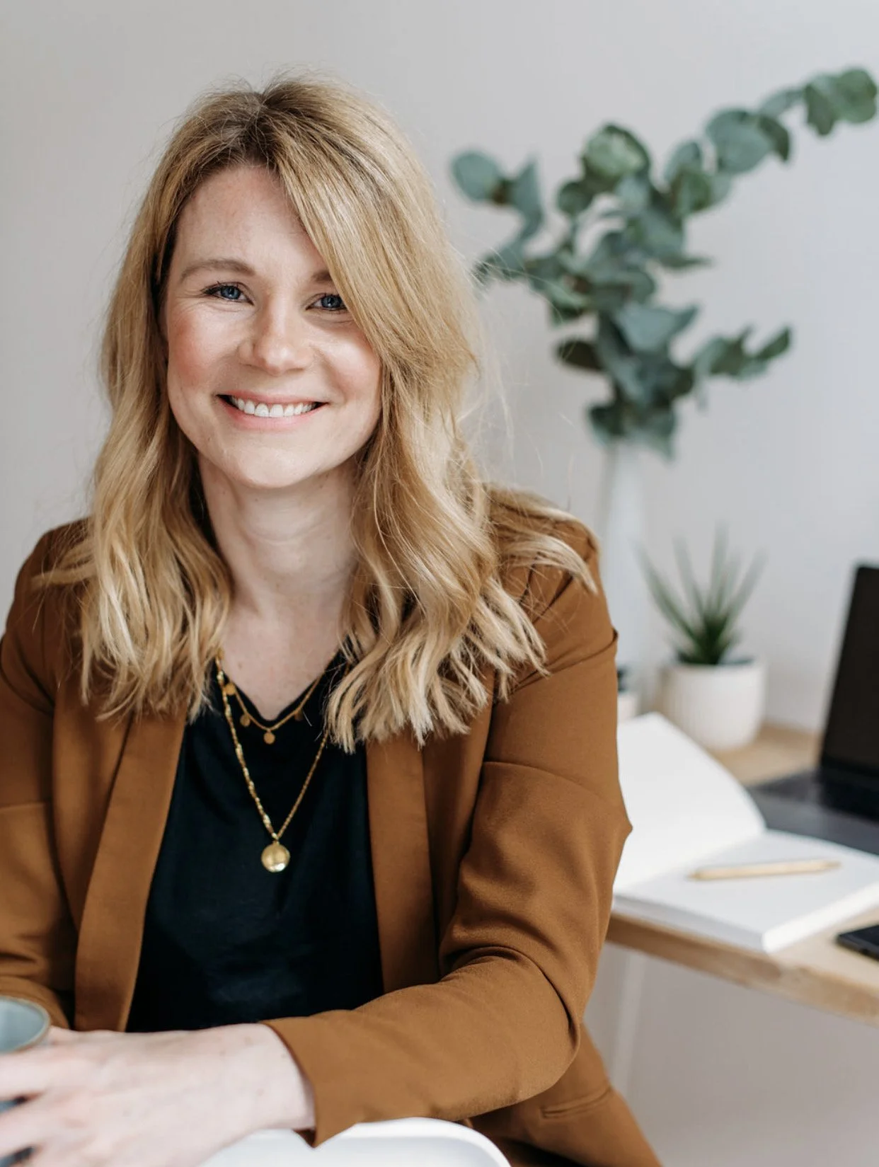 Smiling woman with blonde hair wearing a brown blazer and black shirt, sitting at a desk with a plant and office supplies in the background.