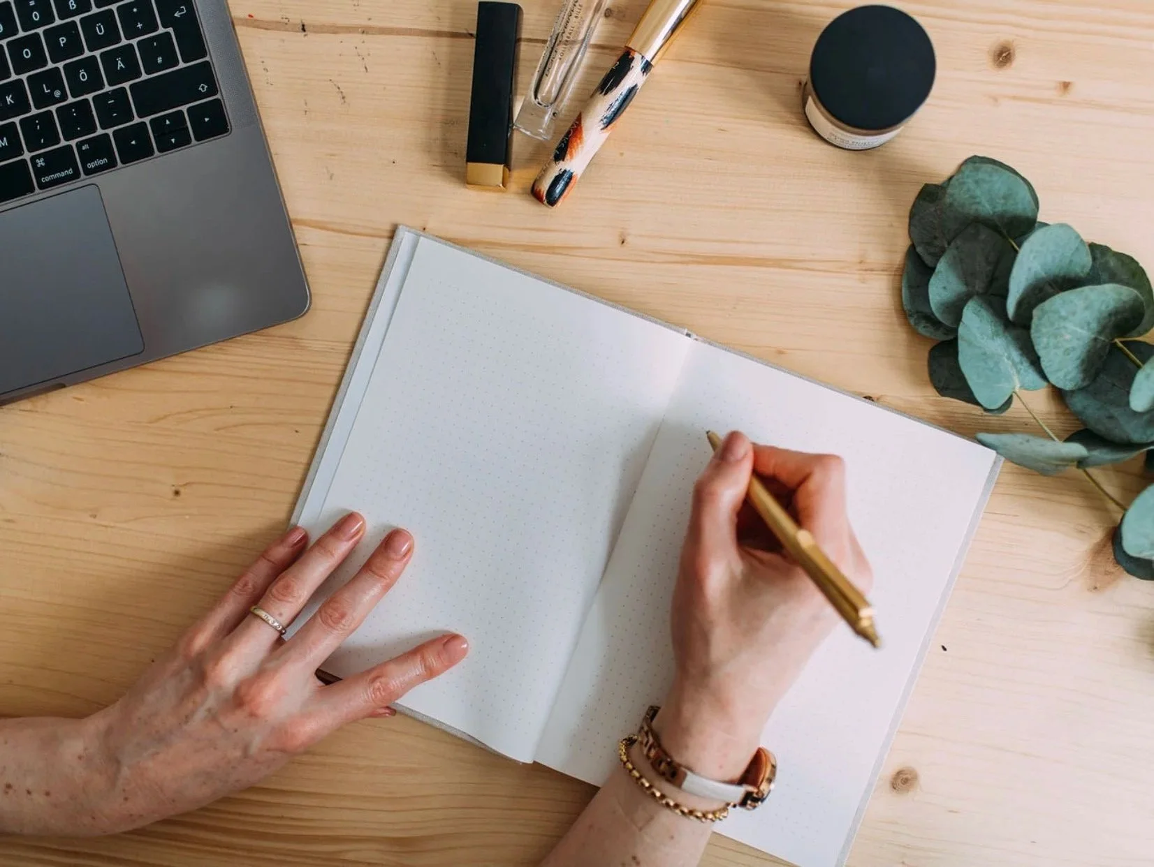 A person writing in a dotted notebook on a wooden desk, with parts of a laptop, pens, a small jar, and green leaves visible on the desk.