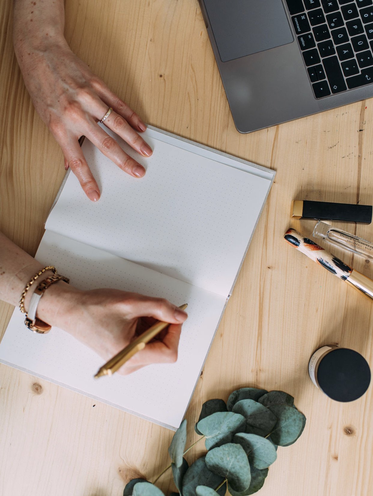 Hands writing in a dotted notebook on a wooden desk with a laptop, pens, a small jar, a black container, and a green leafy plant.