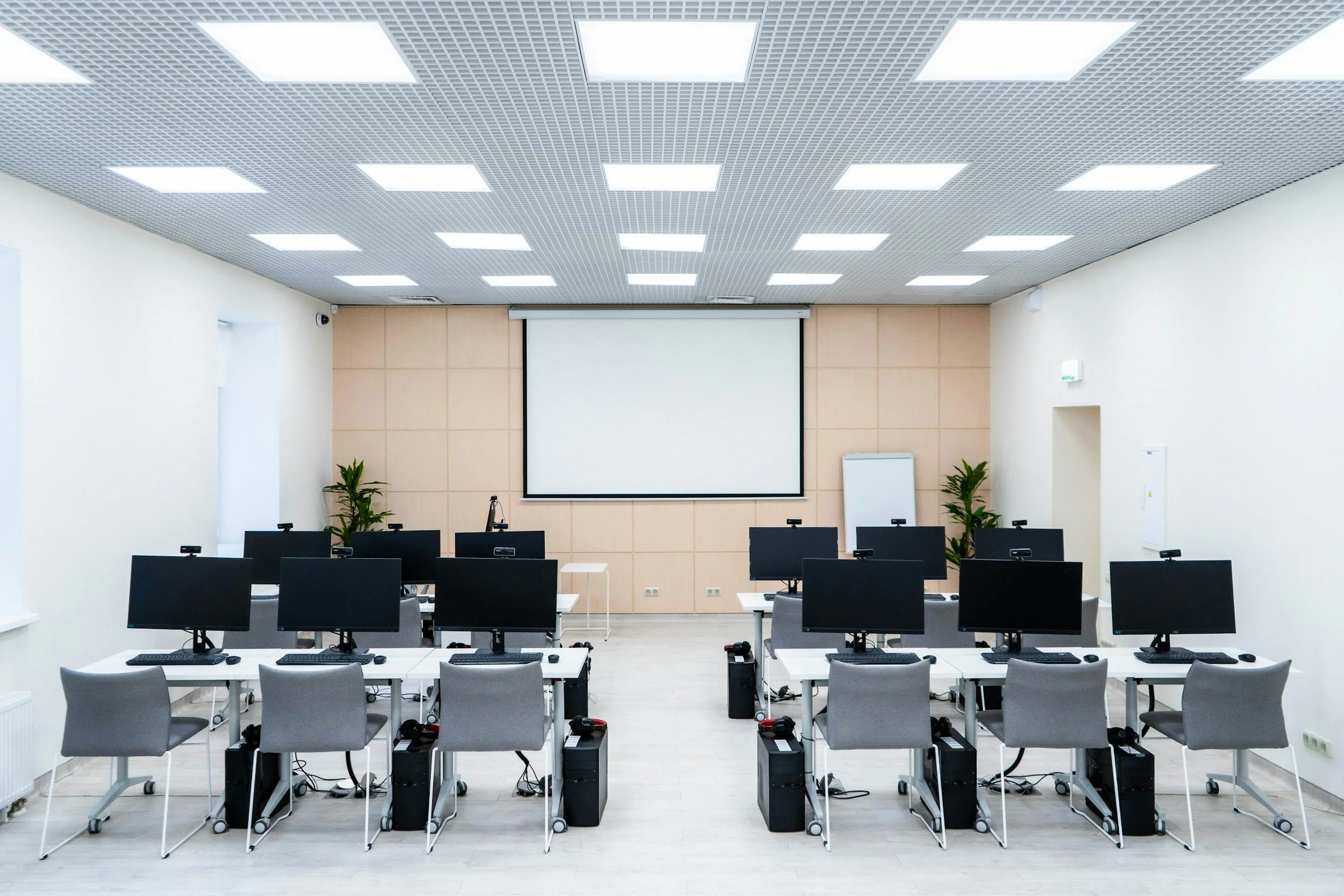 Empty computer training room with rows of desks and monitors, a large screen at the front, and potted plants.