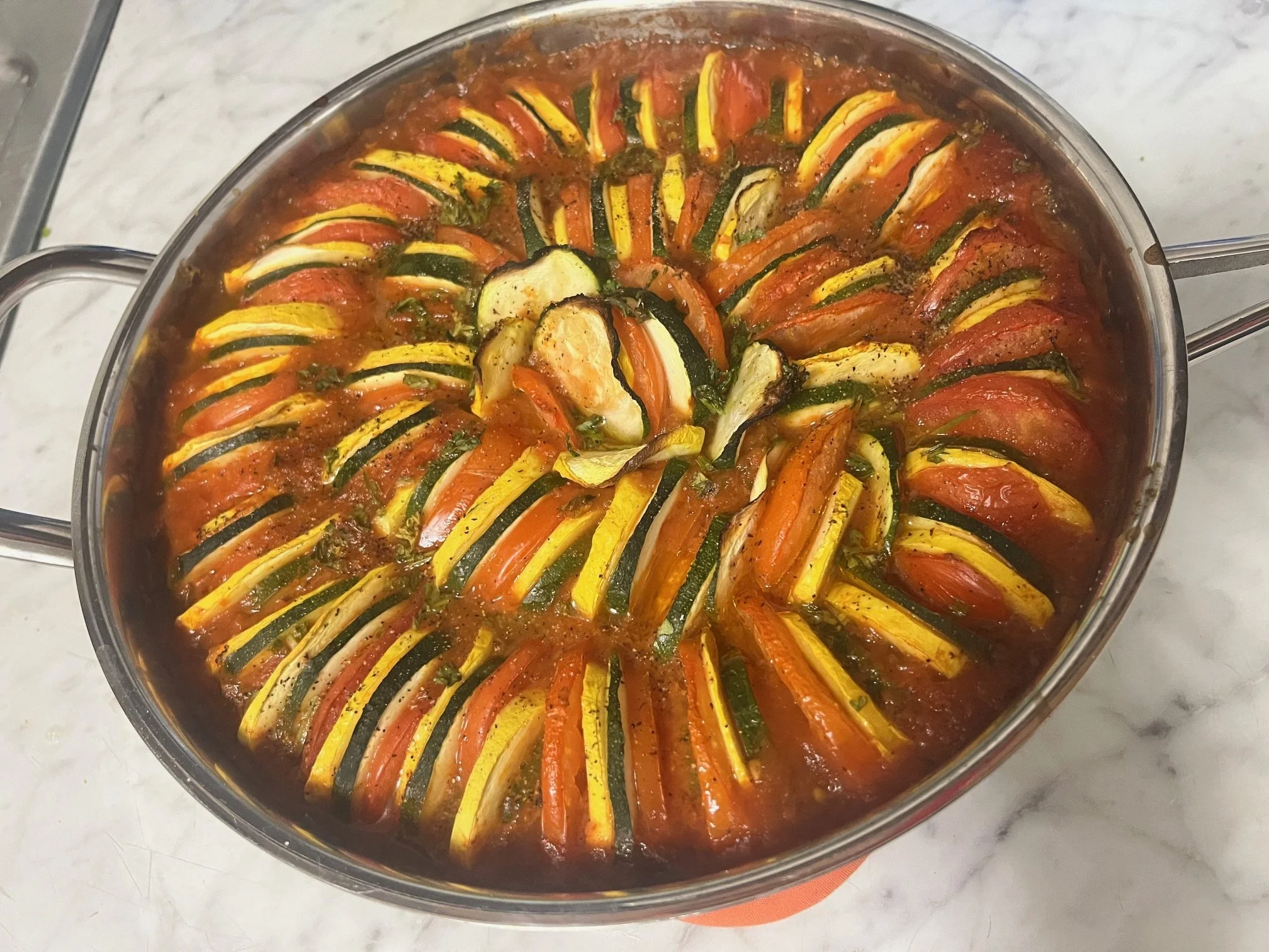 A round glass baking dish containing a dish with alternating slices of yellow squash, zucchini, tomatoes, and carrots arranged in a spiral pattern, cooked in a tomato sauce.