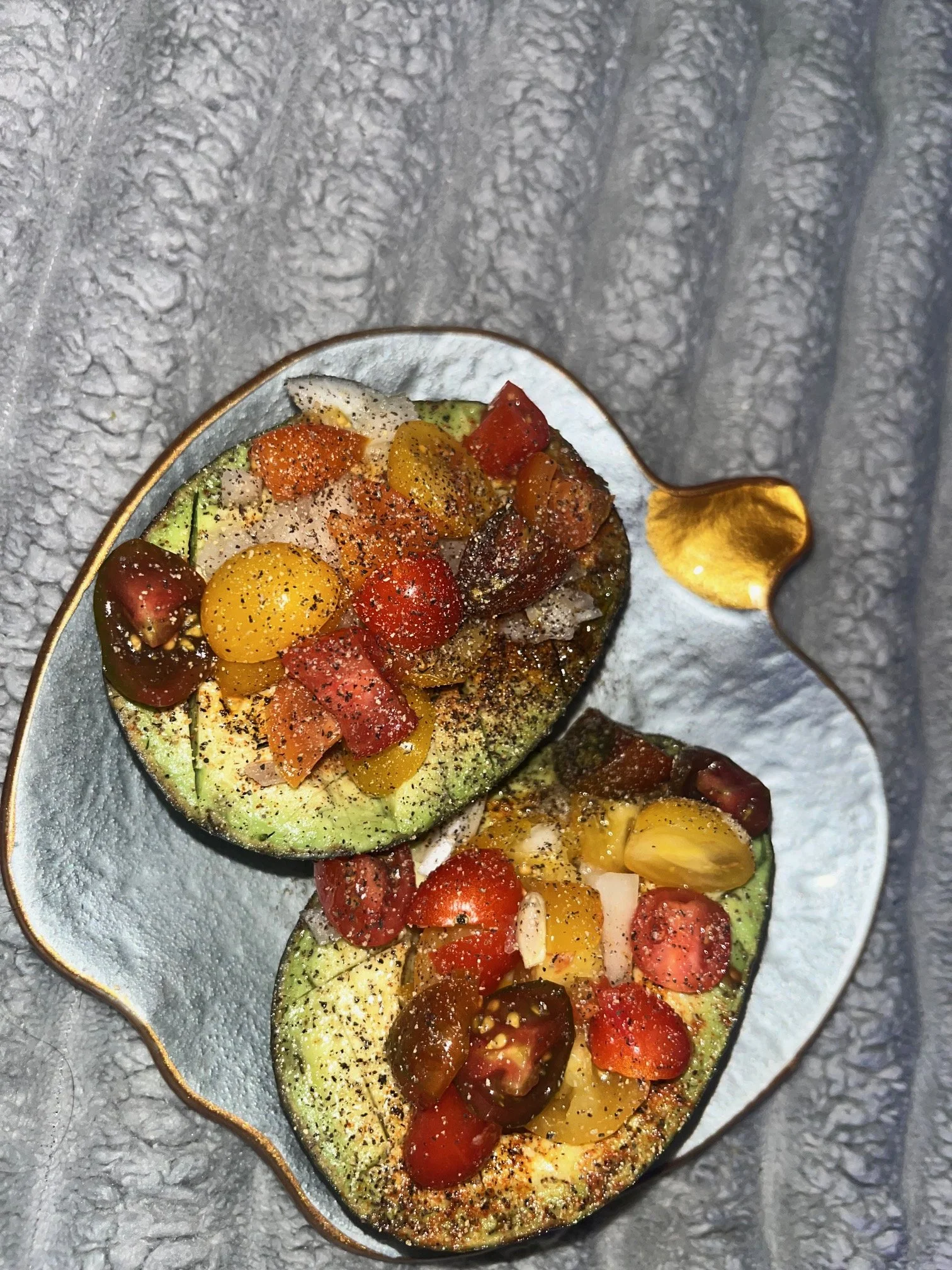 Two avocado halves topped with cherry tomatoes, seasoned with black pepper, served on a decorative plate with a gold handle.