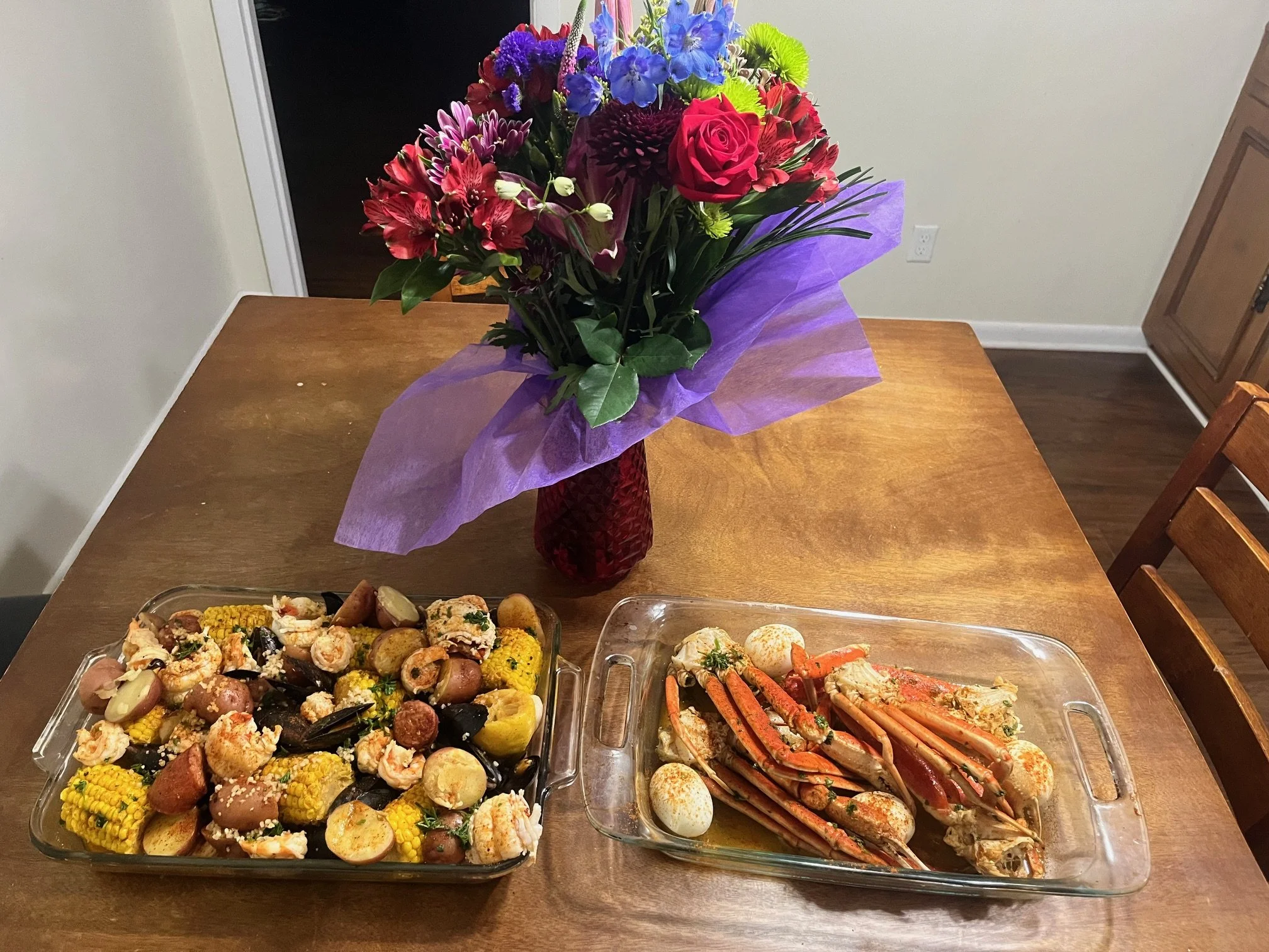 A bouquet of mixed flowers in a red vase, topped with purple tissue paper, placed on a wooden table. Two glass dishes contain cooked seafood and vegetables, including crab legs, potatoes, corn, and sausage, with a slice of lemon visible in the seafood dish.