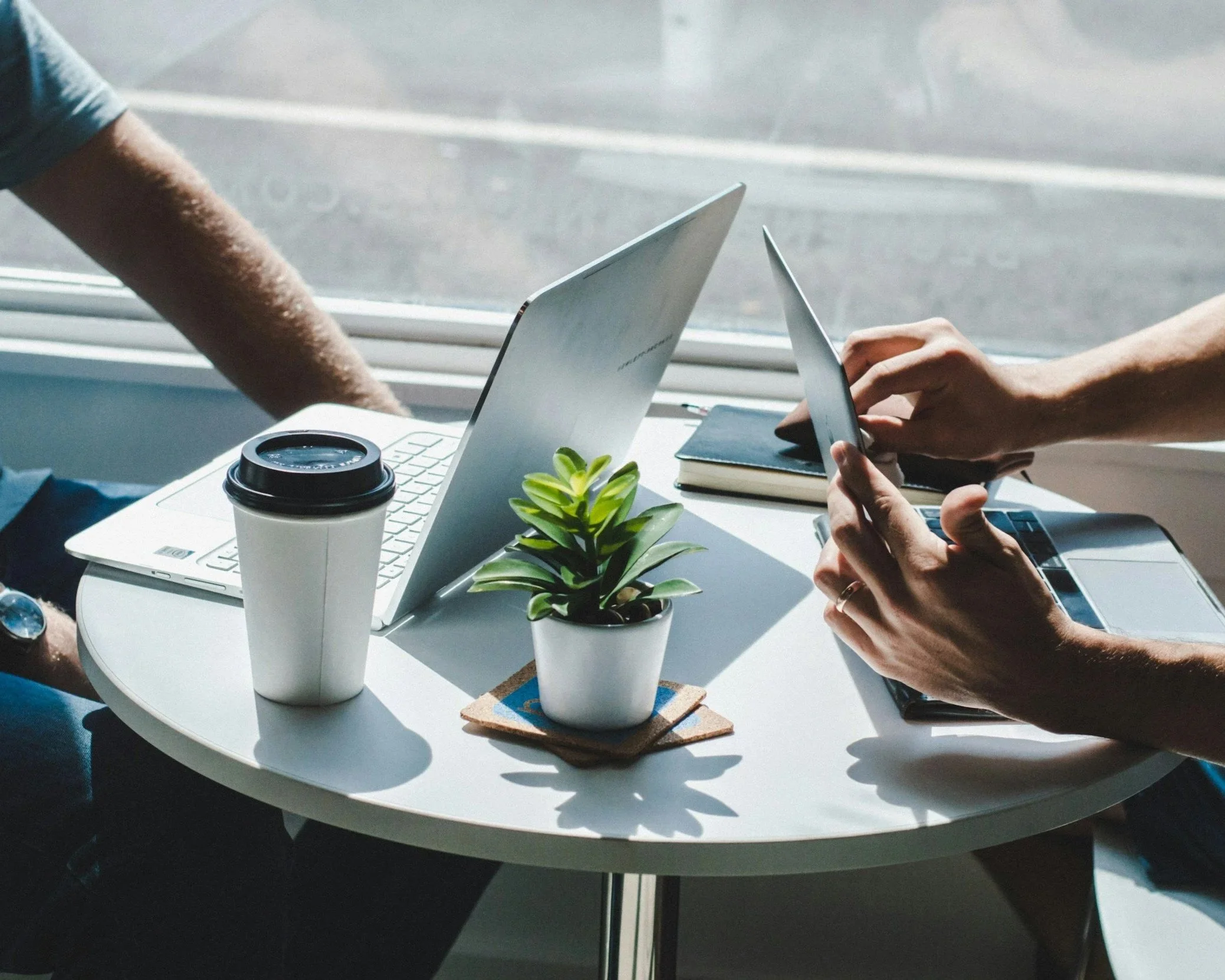 People working at a white table with laptops, a smartphone, a notebook, a cup of coffee or tea, and a small potted plant. Sunlight streams through a window.