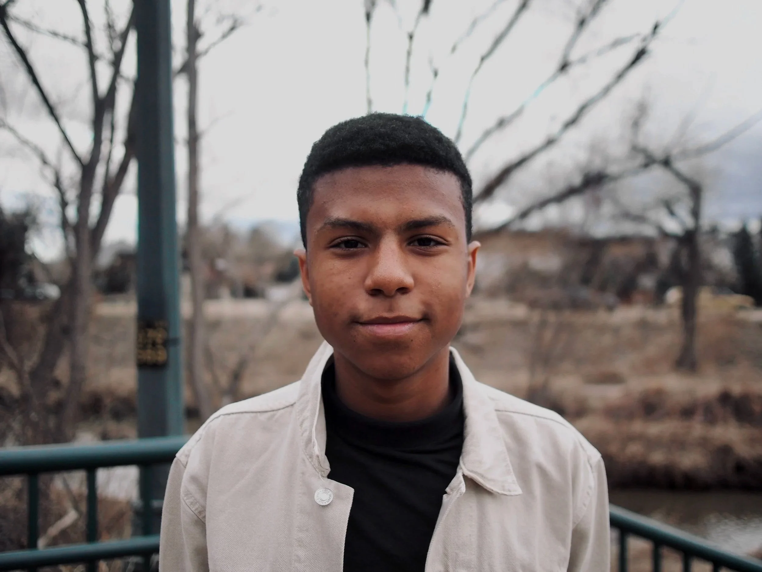 A young man standing outdoors on a bridge with trees and a road in the background.