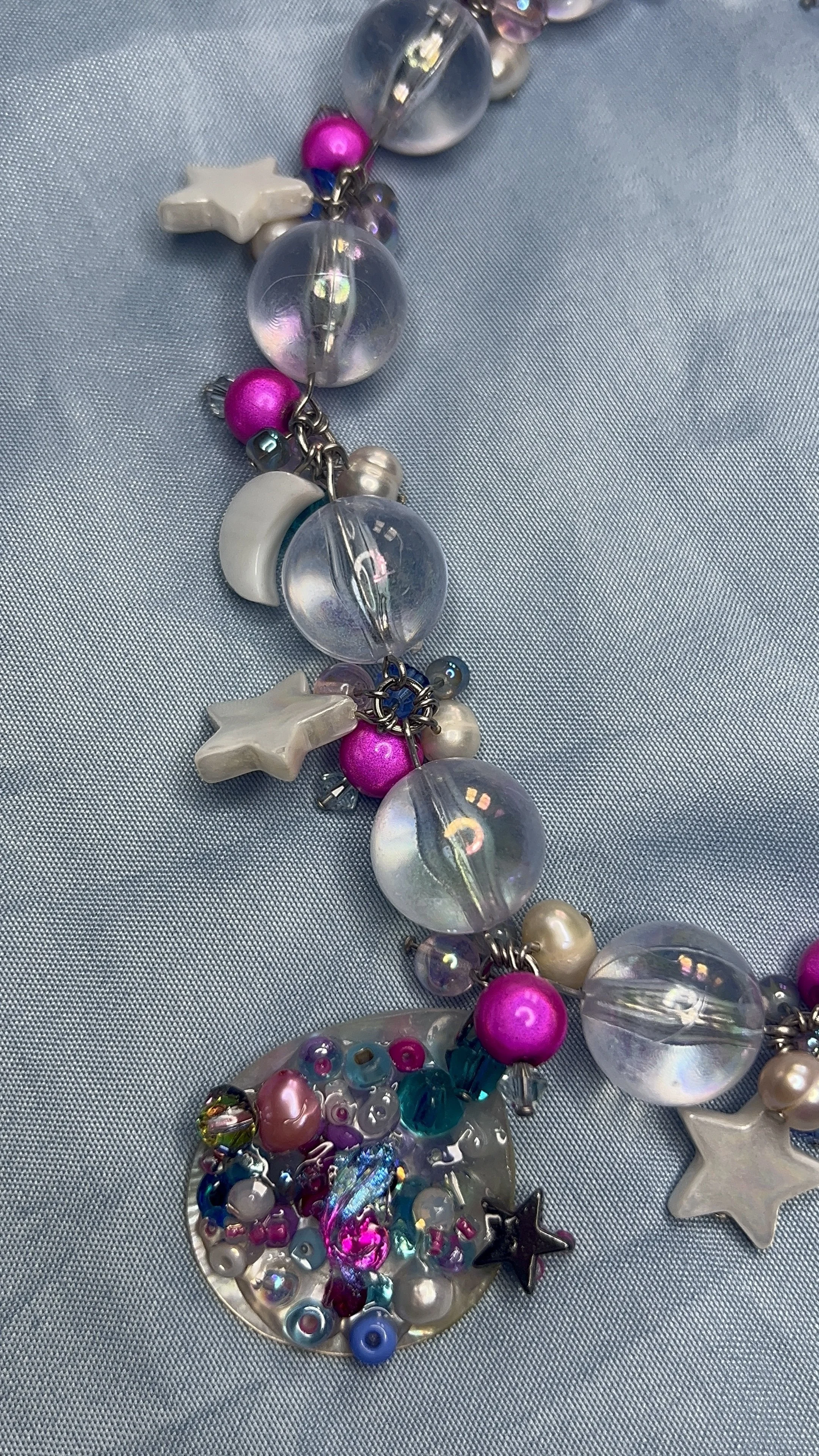 Close-up of a colorful beaded necklace featuring transparent, white, pink, and dark beads, moon and star-shaped charms, and a shell with a "collage" of beads on light blue fabric.