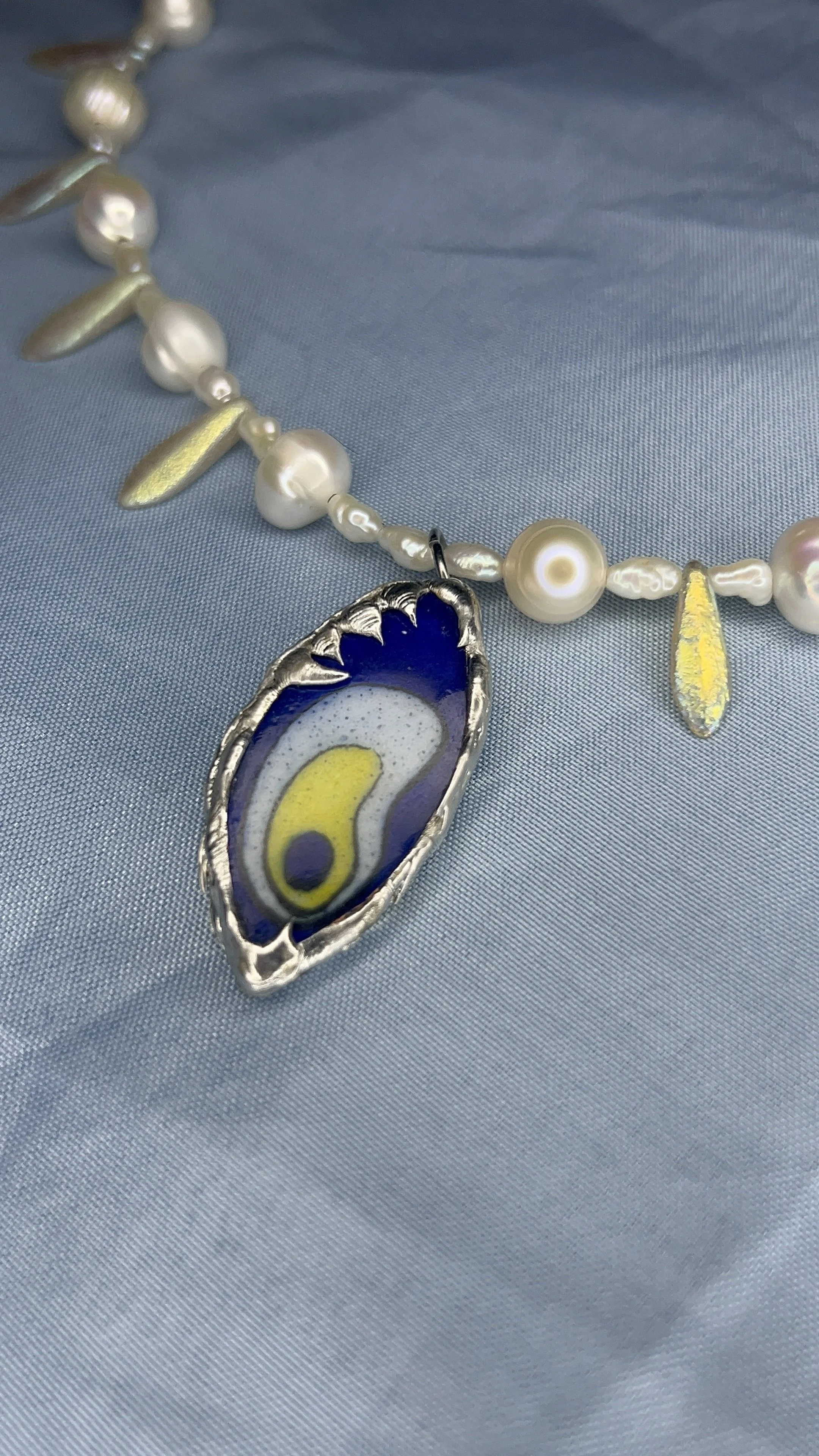 Close-up of a pearl necklace with gold accents and an enamel pendant on a light blue fabric background.