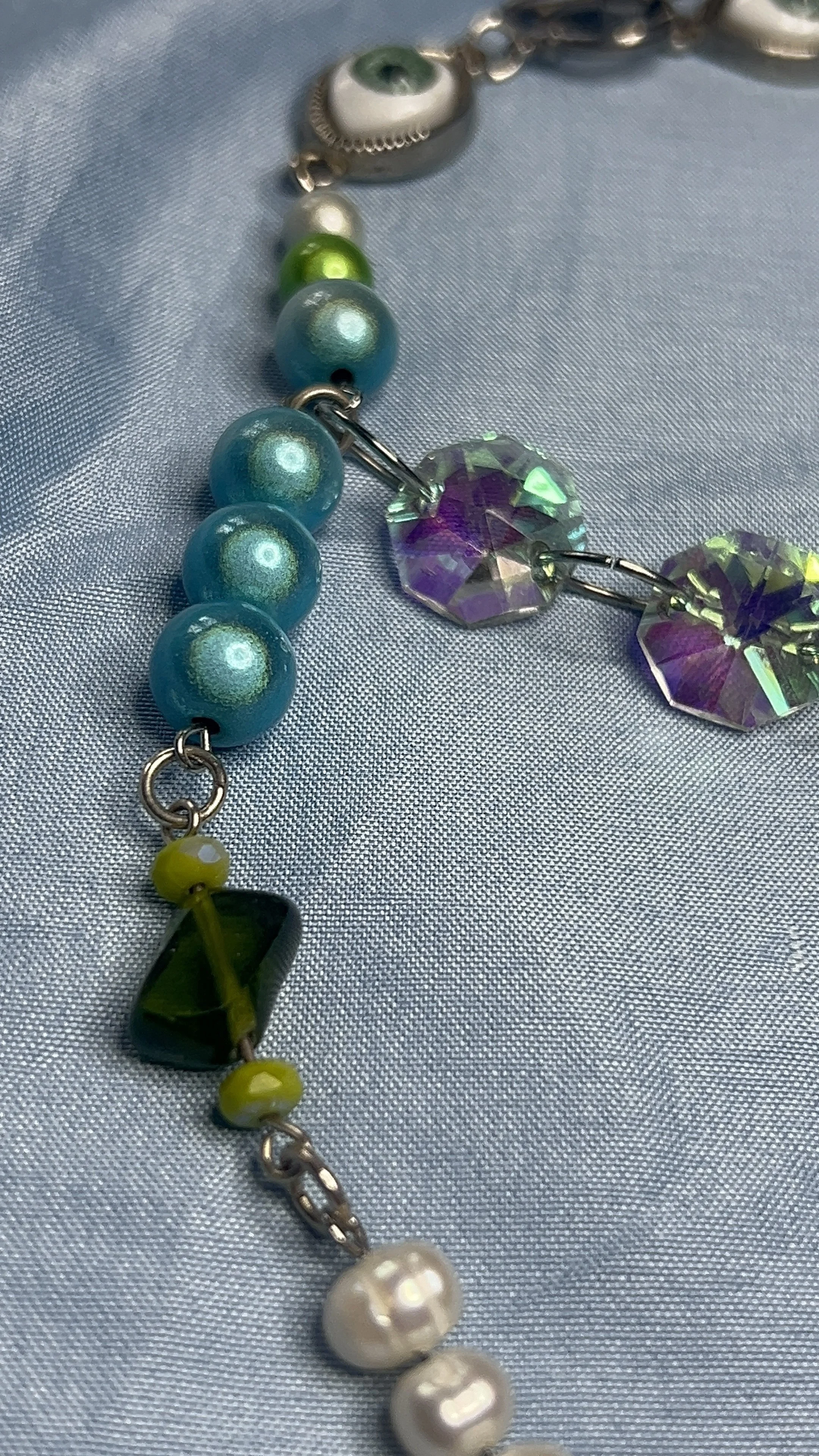 Close-up of a colorful beaded necklace with beads in shades of blue, green, purple, and white, laid on light blue fabric.