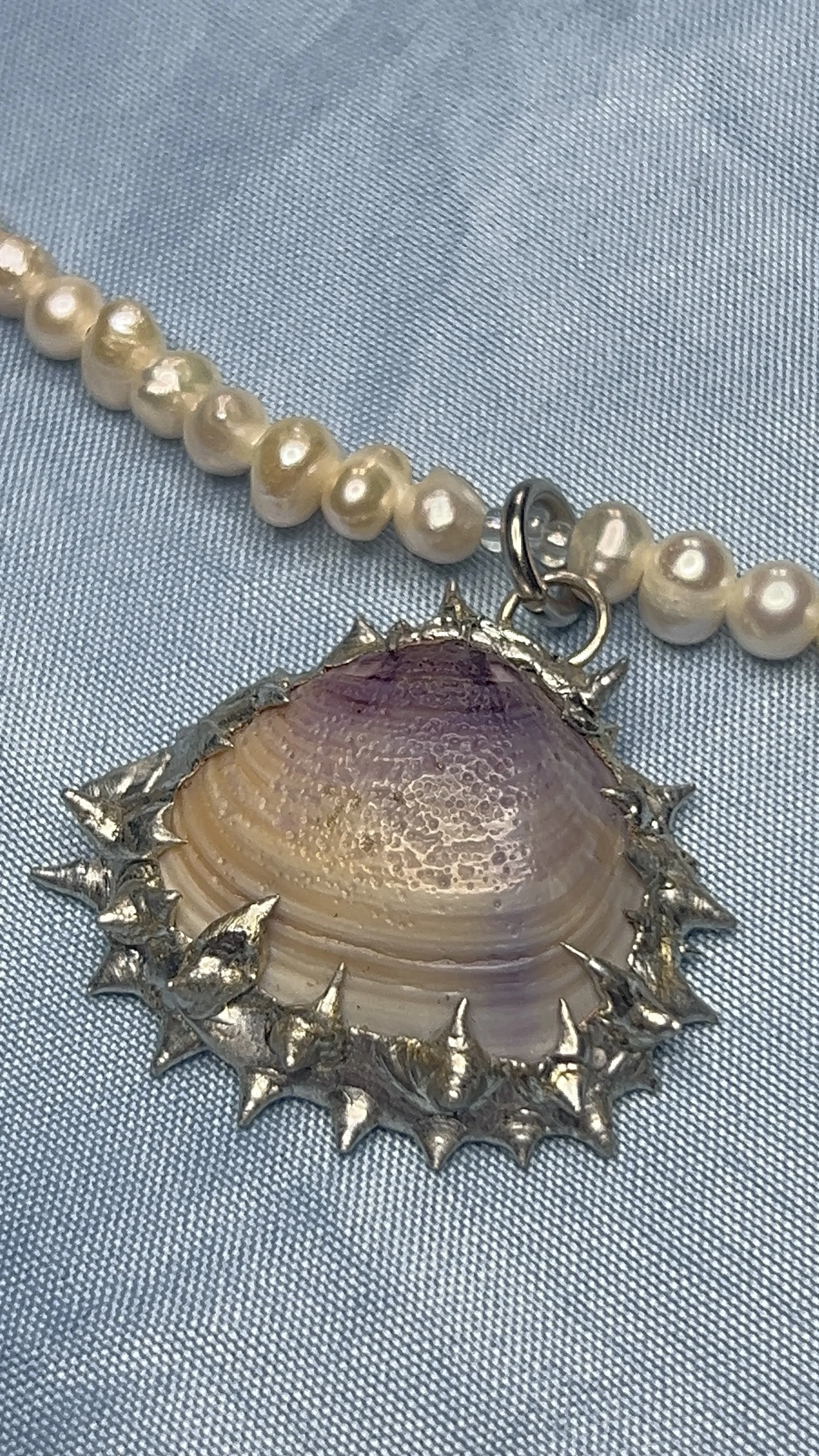 A pearl necklace with a large shell pendant surrounded by a spiked metal frame, placed on blue fabric.