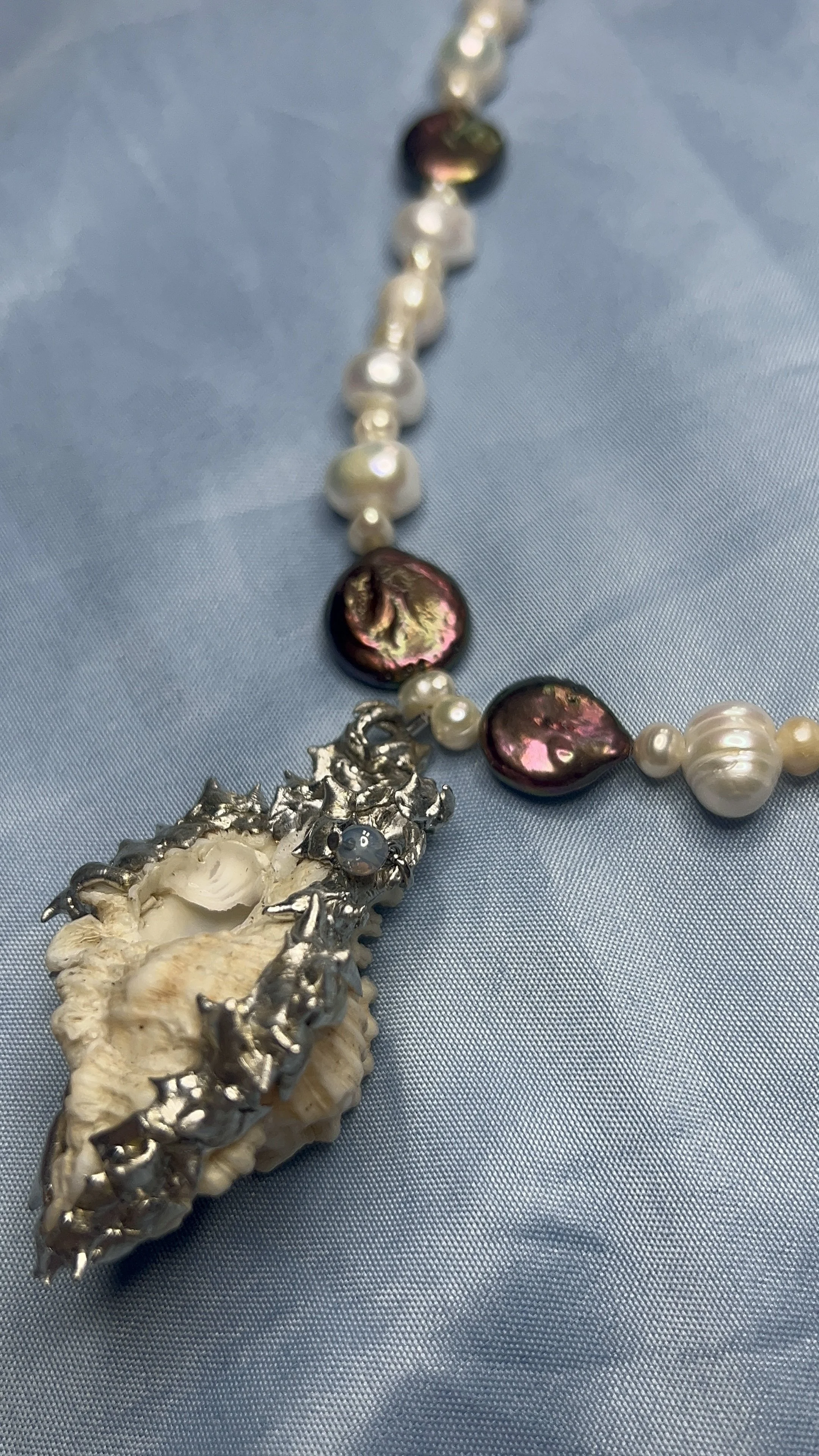 A close-up of a necklace with pearls, iridescent stones, and a large shell pendant with silver accents on a light blue fabric background.