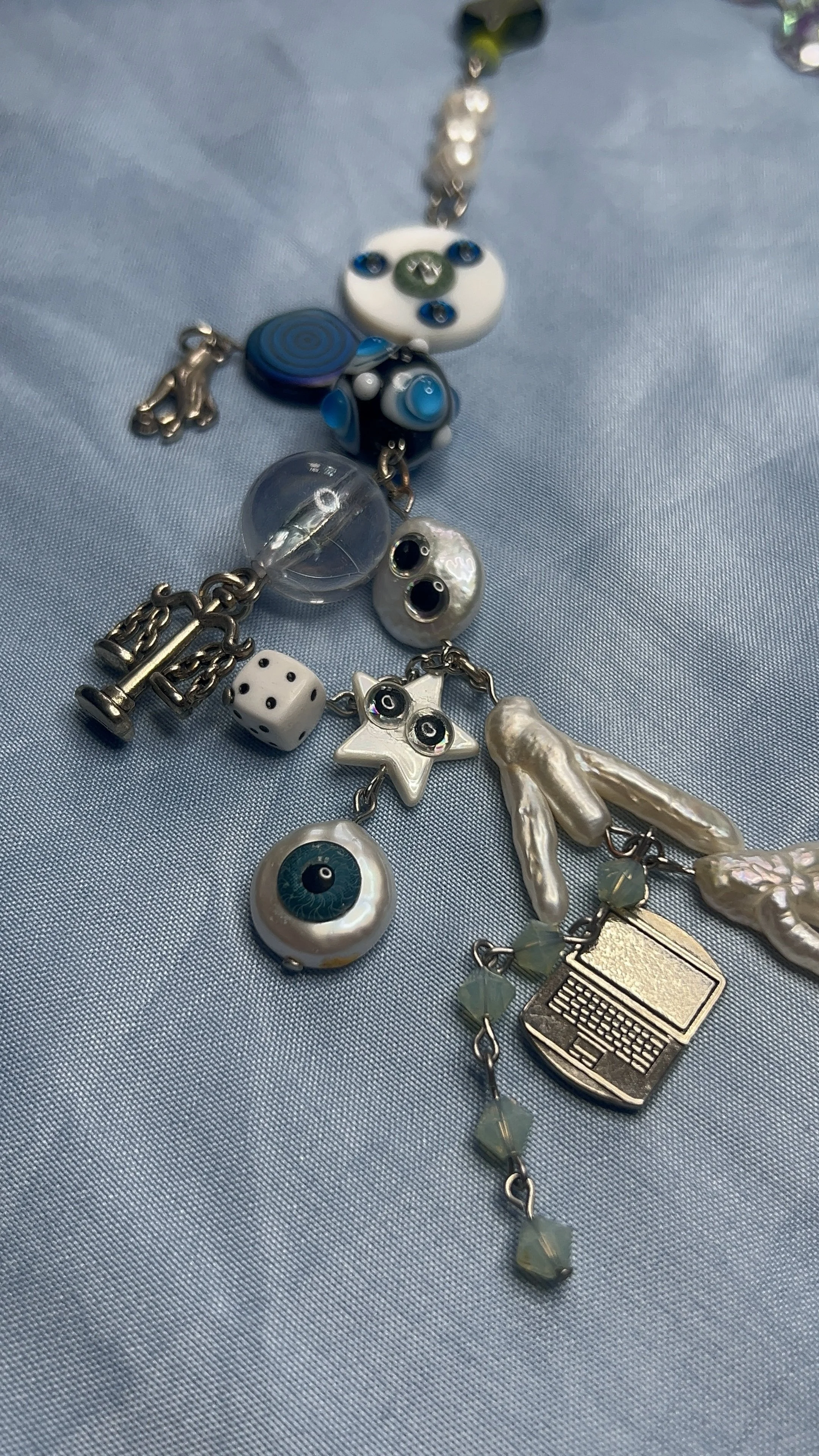 A collection of various charms and pendants on a light blue fabric surface, including an eye, a star with eyes, a laptop, a die, and several pearl-like and glass beads.