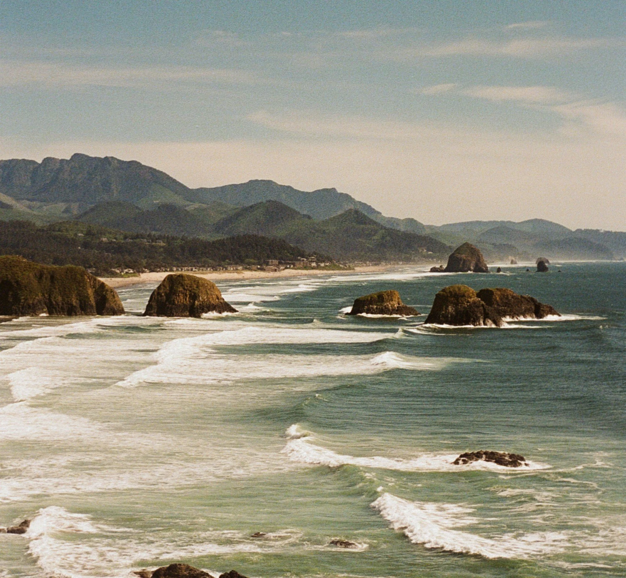 View of a rugged coastline with large sea stacks, sandy beach, and green mountains in the background under a cloudy sky.