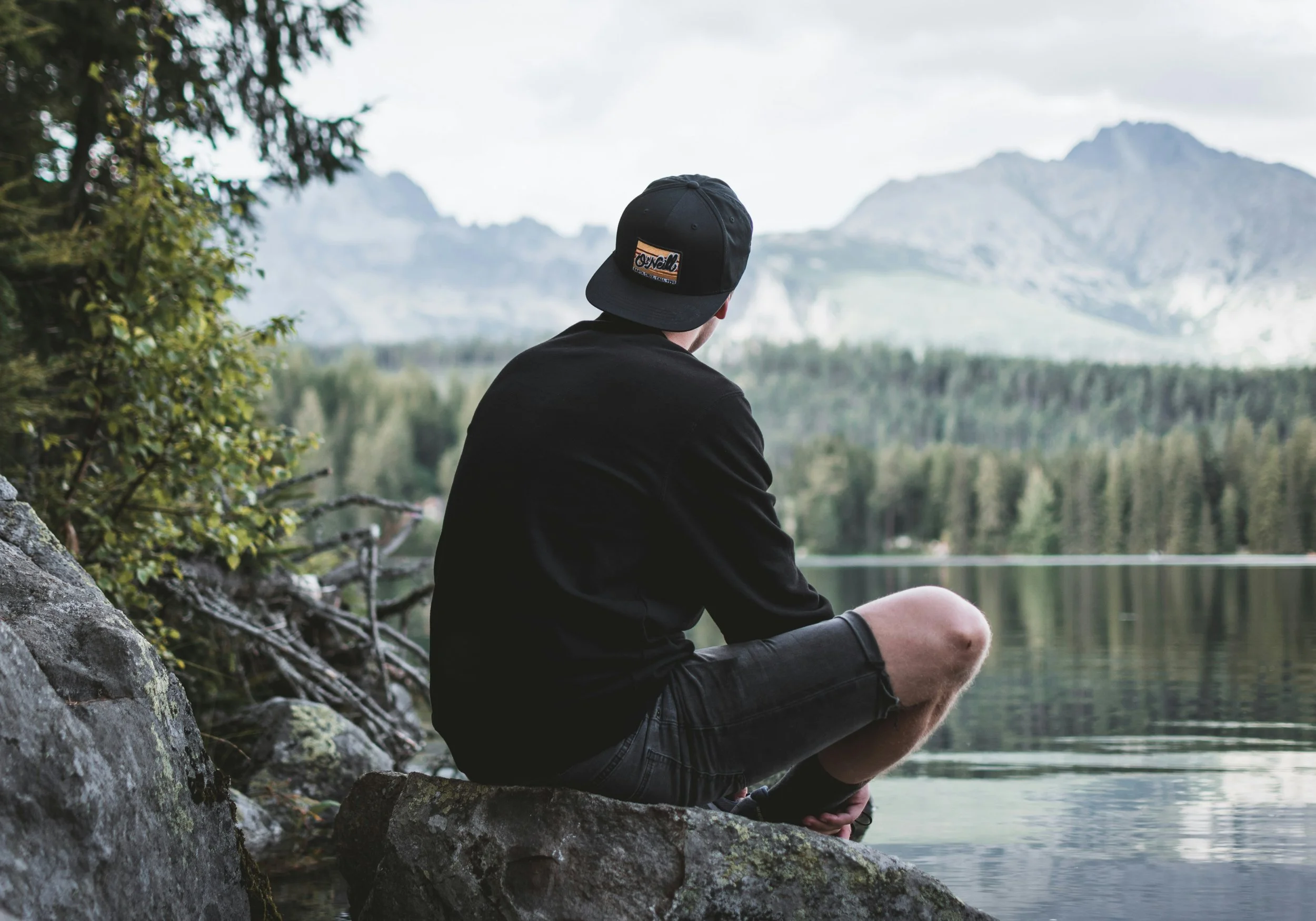 Person sitting on rock by lake, looking at mountains and forest, wearing black hoodie, shorts, and a black cap.