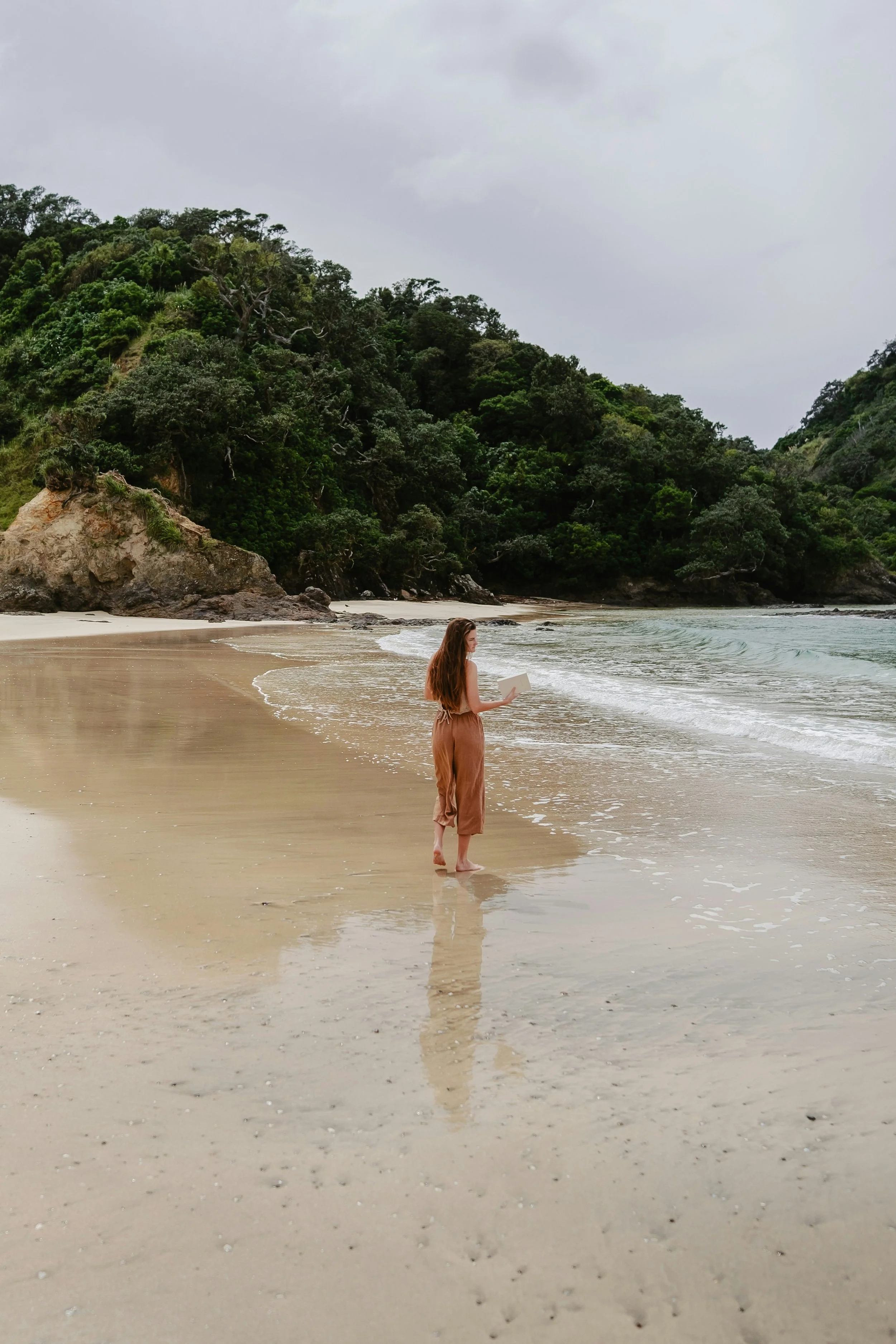 A woman with long brown hair stands barefoot on a sandy beach, holding a laptop, with waves and a forested hillside in the background under a cloudy sky.
