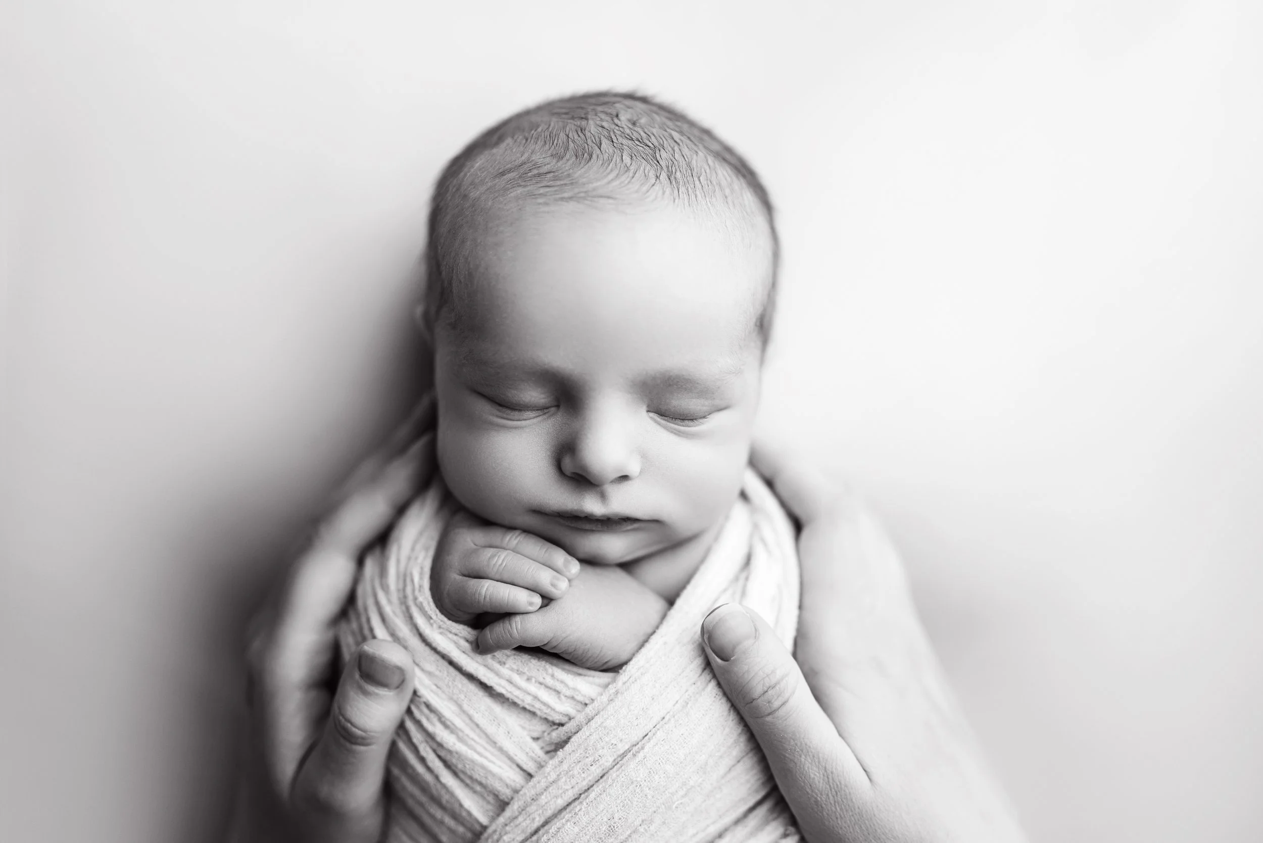 Black-and-white photograph of a sleeping Calgary newborn baby wrapped in a cloth, gently held by an adult's hands at their calgary newborn session