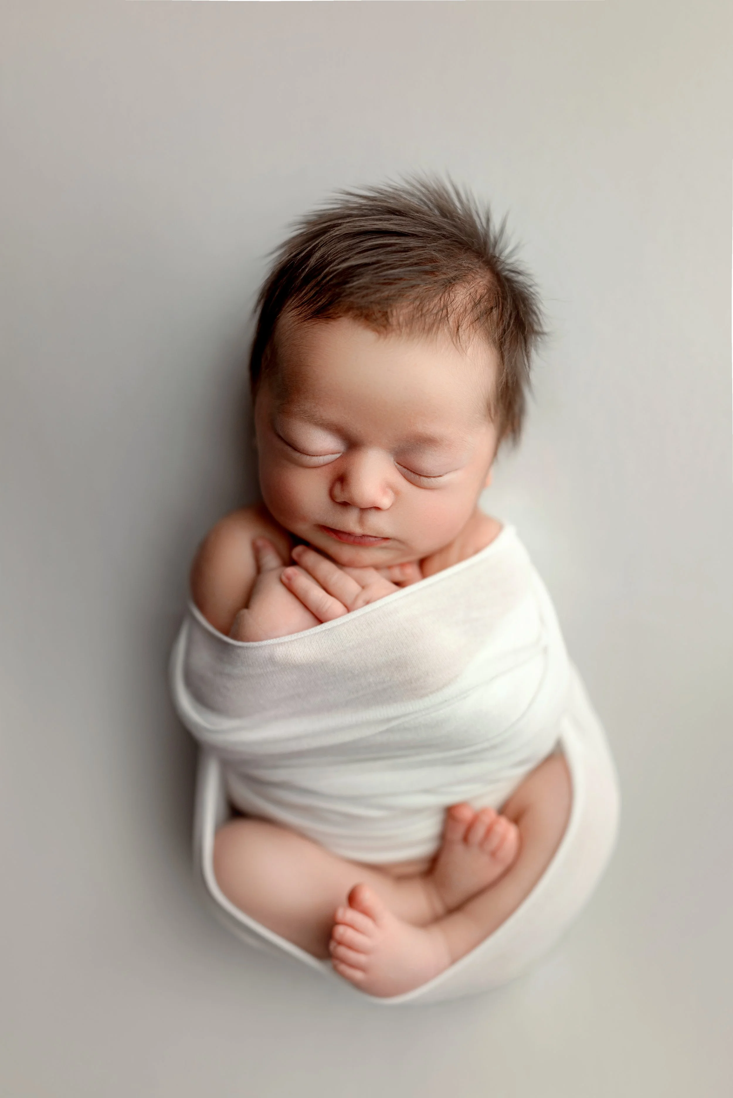 Calgary newborn session of a sleeping baby wrapped in a white blanket with hands resting near their chin, lying on a light grey surface.