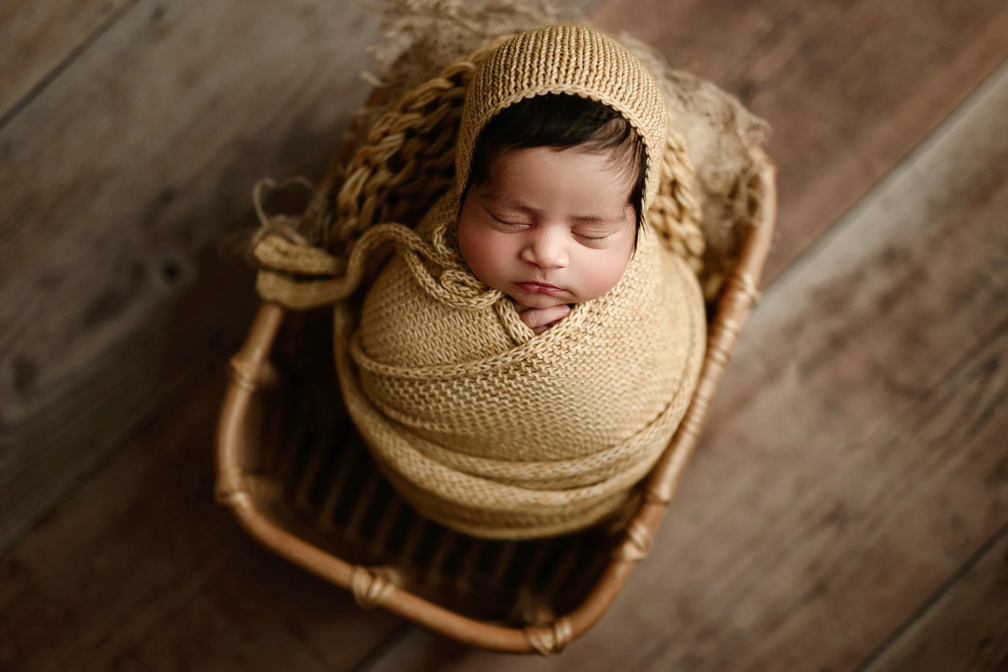 A calgary newborn baby sleeping at their newborn session wrapped in a beige knitted blanket and wearing a matching cap, lying in a small woven basket on a wooden floor.
