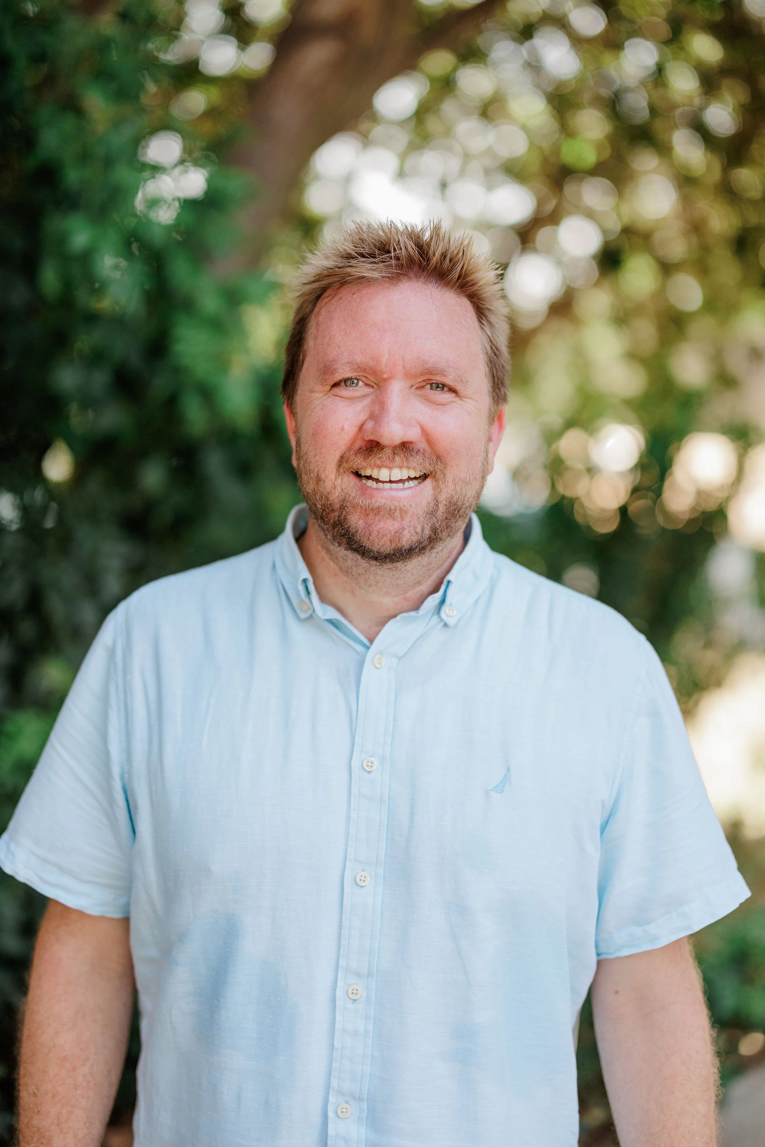 Close-up photo of a smiling man with light hair and a beard, wearing a light blue shirt, standing in front of an trees outdoors