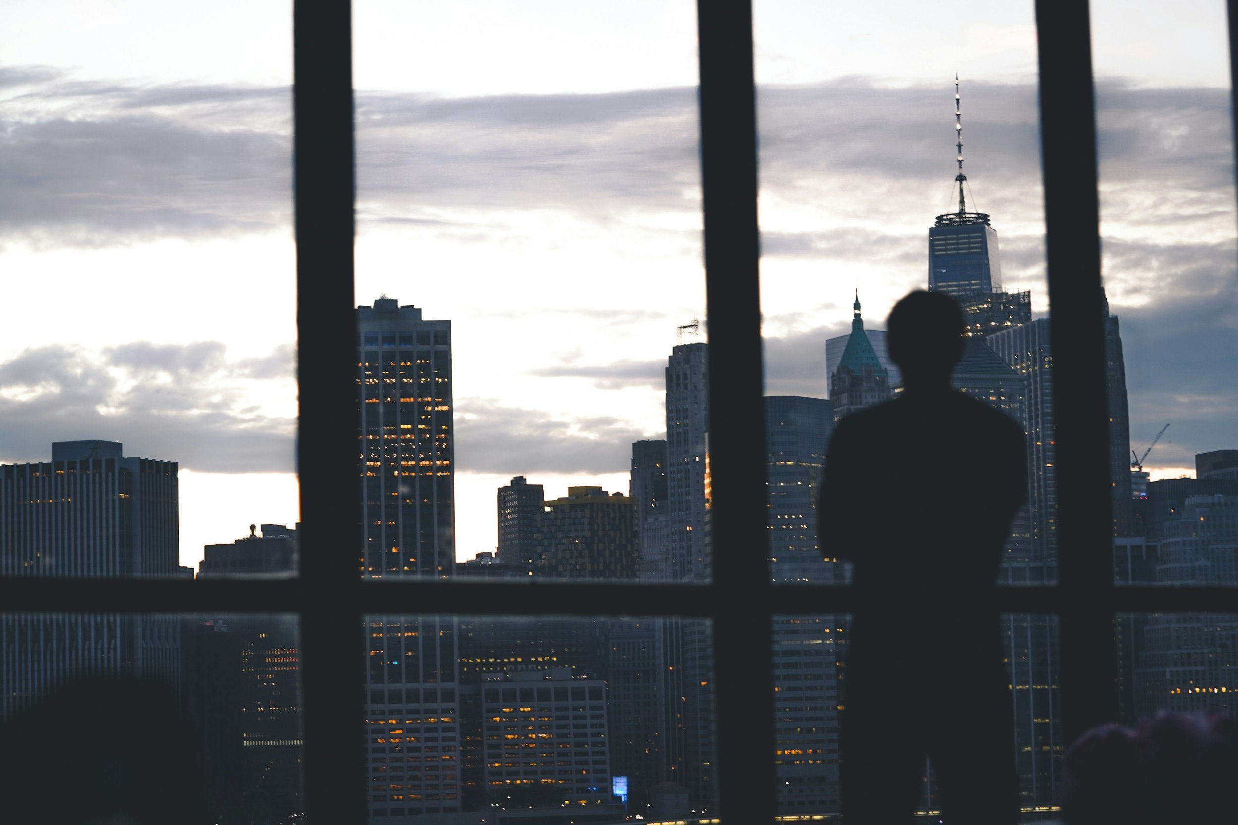 A silhouette of a person observing a city skyline through window bars, with tall buildings including the One World Trade Center and other skyscrapers under a cloudy sky.