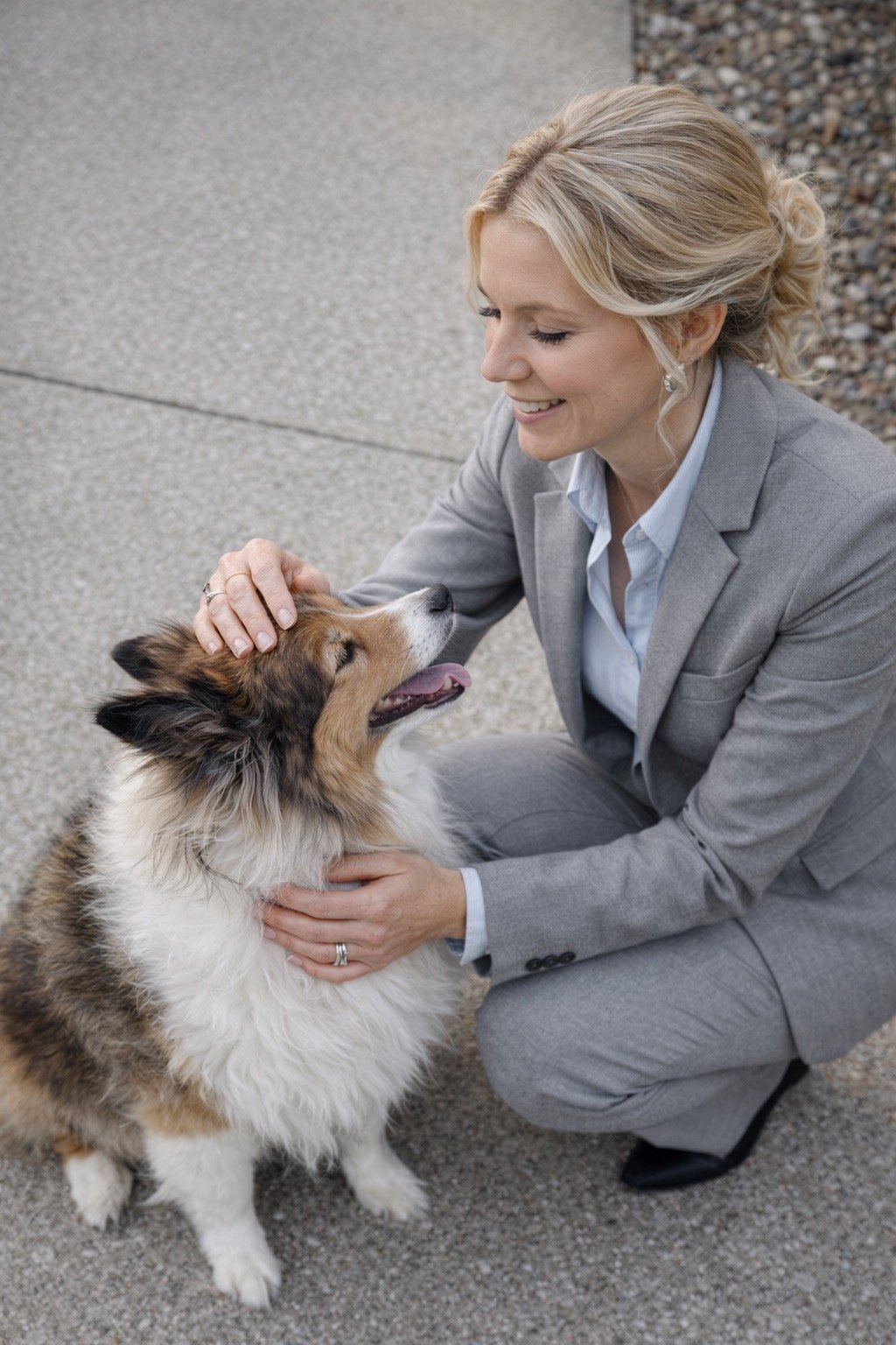 A woman in a gray suit crouches down and pets a happy, fluffy Australian Shepherd dog on a sidewalk.