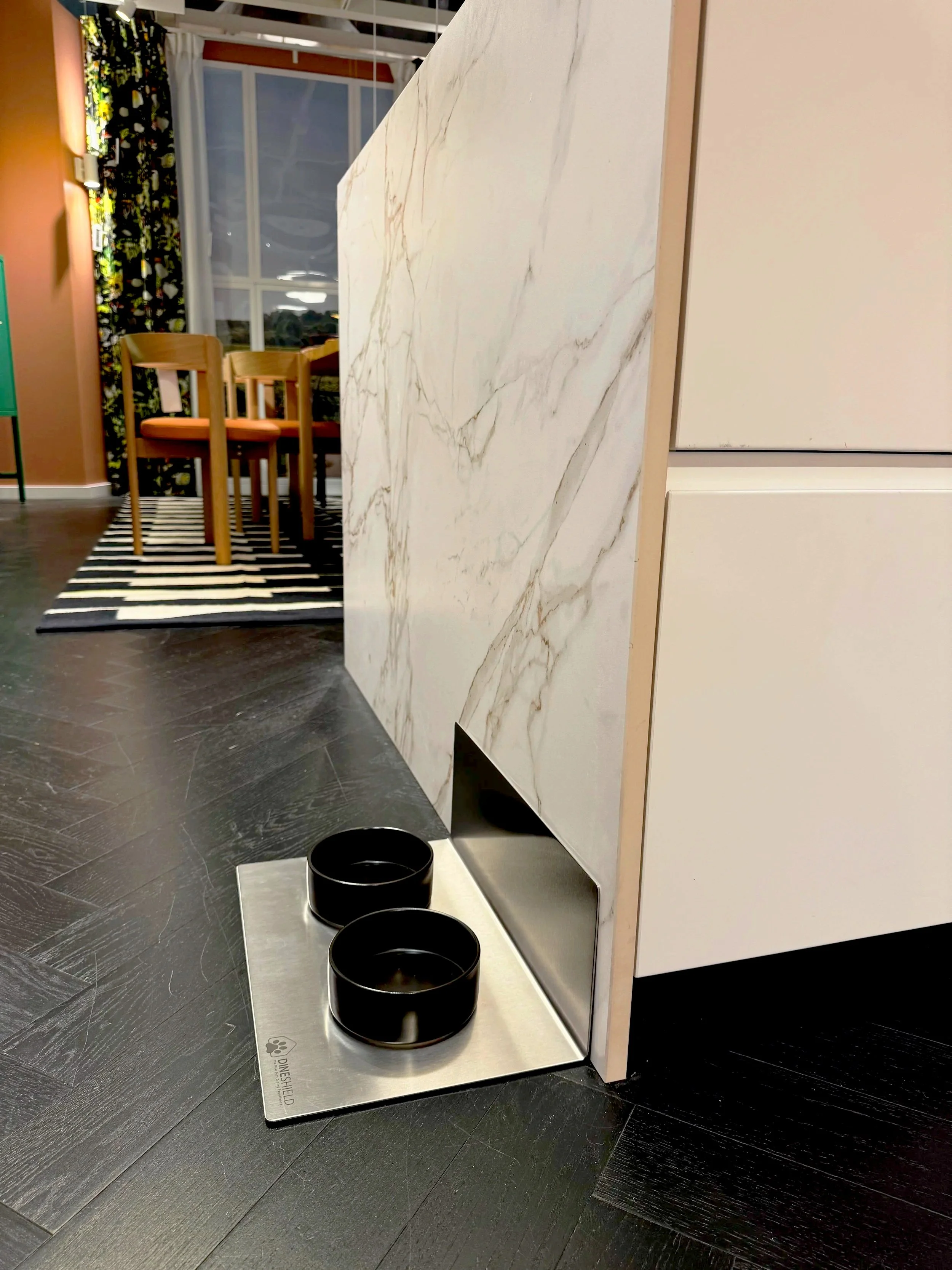 DineShield Edelstahl-Napfunterlage mit schwarzen Näpfen vor weißer Marmorwand auf dunklem Holzboden – modernes Premium-Design