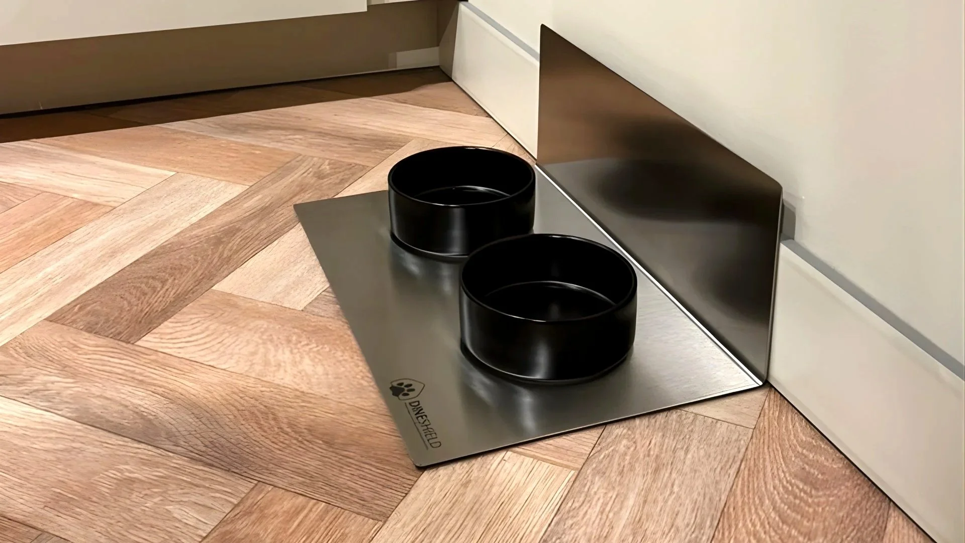 Seitenansicht: DineShield Edelstahl-Napfunterlage mit zwei schwarzen Näpfen auf Fischgrätparkett – L-Form an der Wand