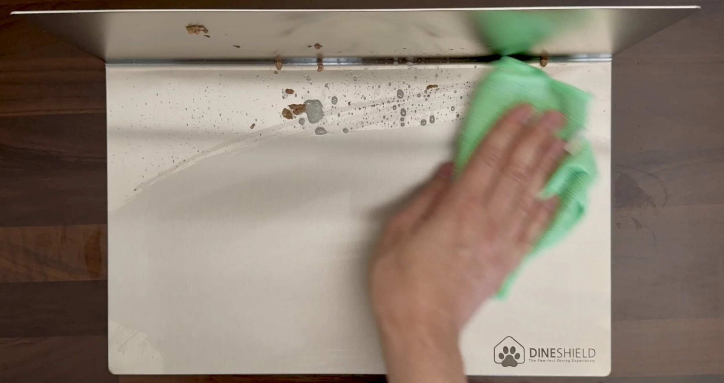 Reinigung von DineShield: Hand wischt Edelstahl-Napfunterlage mit Mikrofasertuch – Schmutz wird sichtbar entfernt