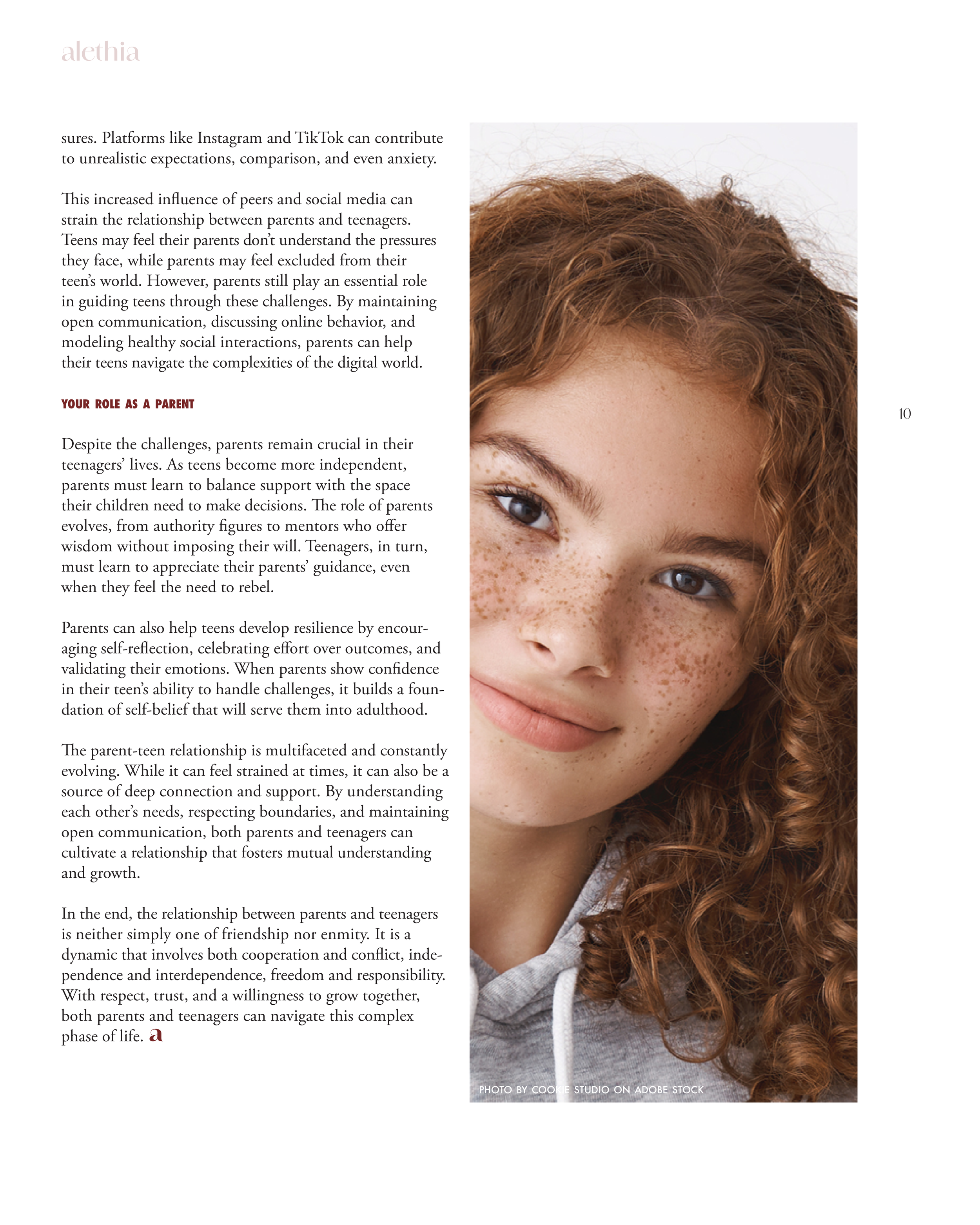 A close-up portrait of a smiling girl with curly red hair, freckles, and light skin, wearing a gray hoodie.