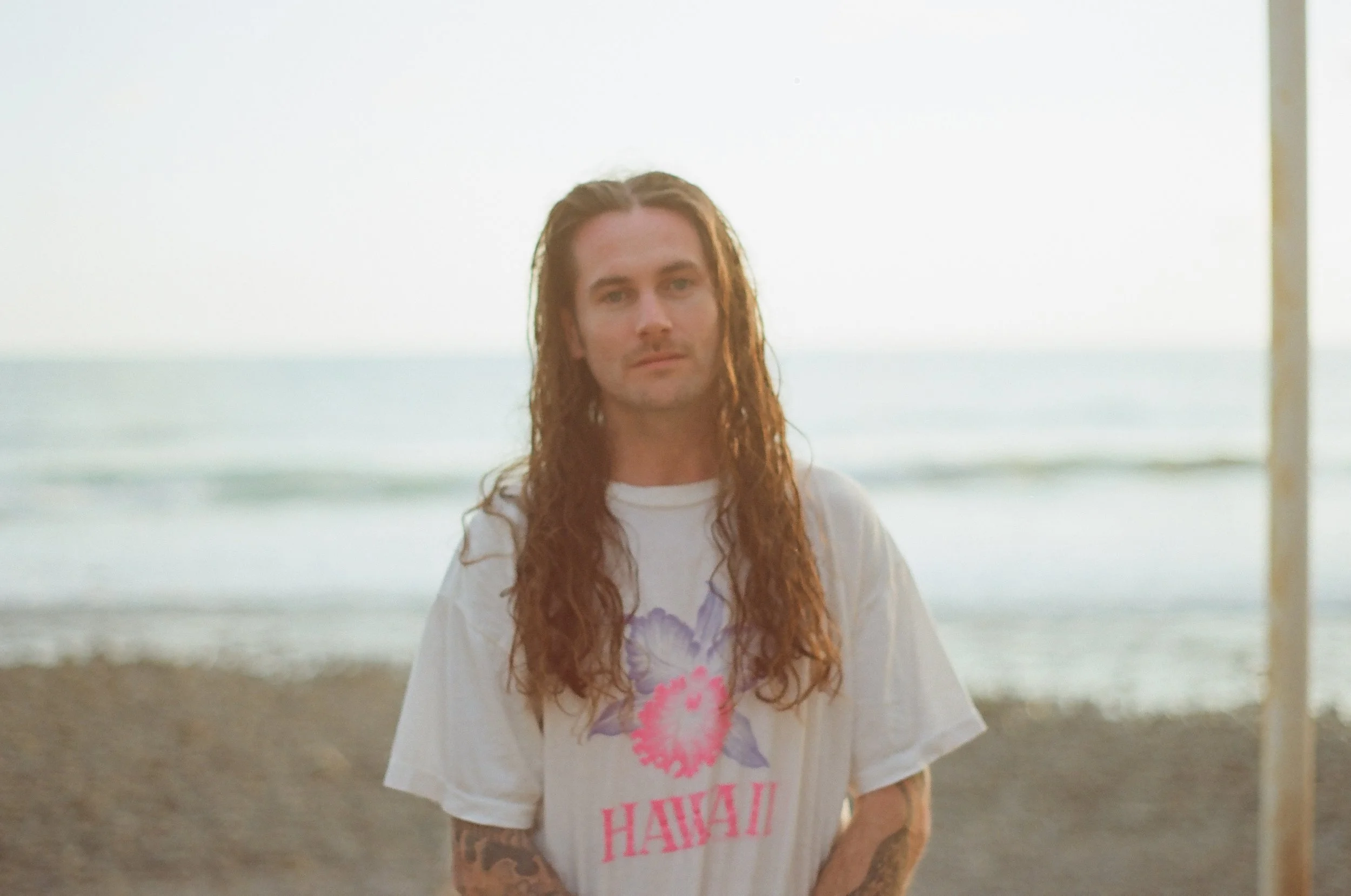 A man with long, wavy hair stands on a beach during sunset, wearing a white T-shirt with a floral design and the word 'Hawaii' printed on it.