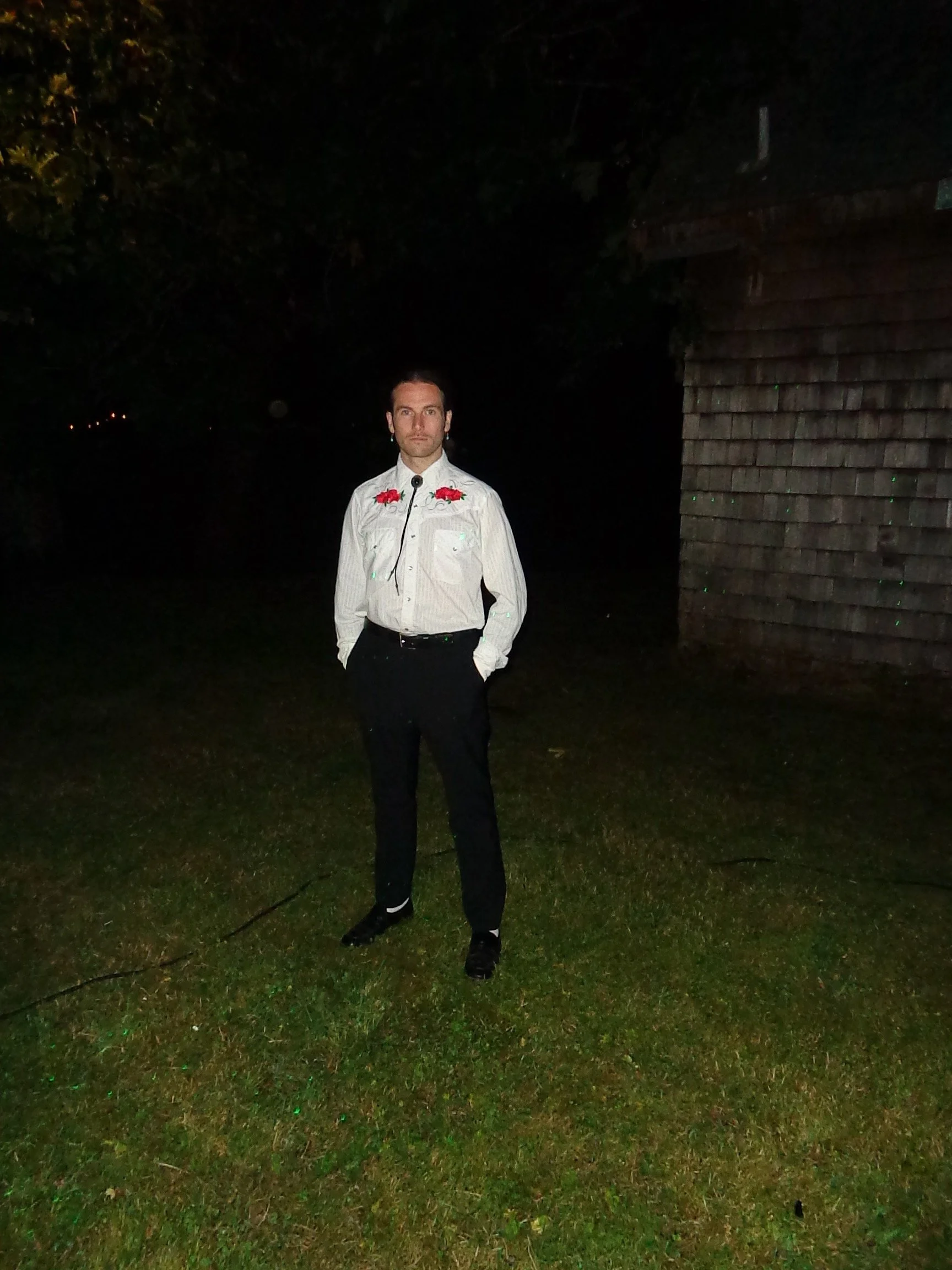 A man standing on grass at night, wearing a white shirt with embroidered roses on the shoulders, black pants, and black shoes, with hands in his pockets and looking at the camera.