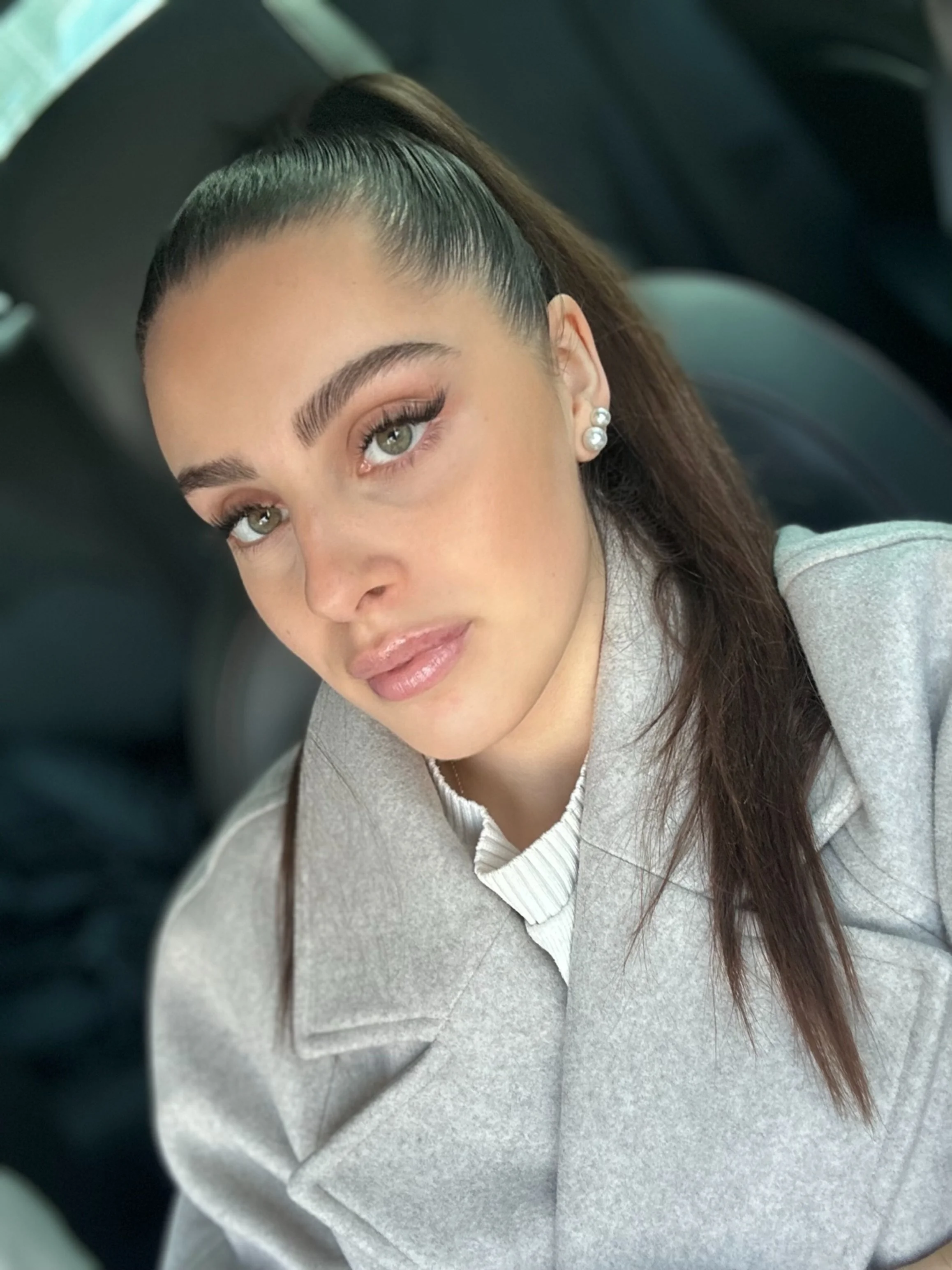 Nahaufnahme einer jungen Frau mit braunen, langen Haaren, die zu einem Pferdeschwanz gebunden sind, und mit Make-up, die in einem Auto sitzt, trägt Ohrringe und eine graue Jacke.
