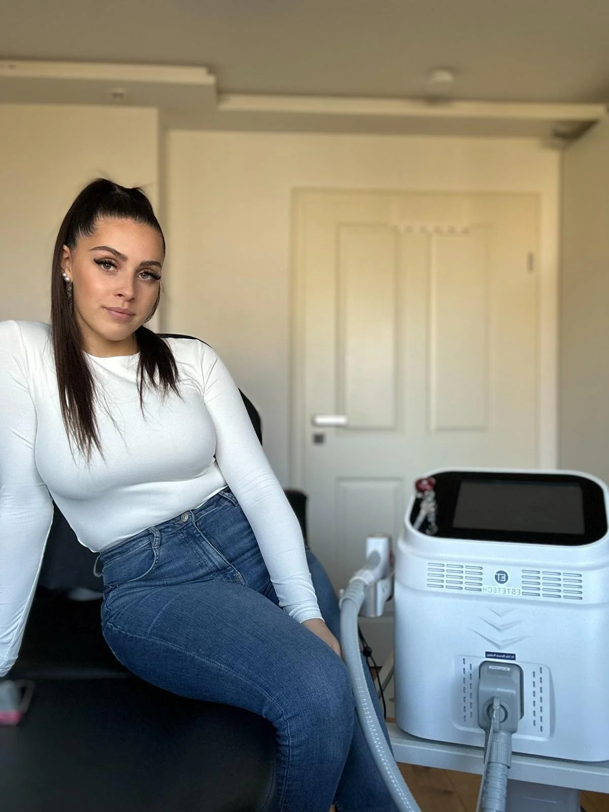 Junge Frau sitzt auf einem schwarzen Stuhl in einem Raum mit weißen Wänden, neben einer medizinischen Maschine, vermutlich bei einer kosmetischen Behandlung.