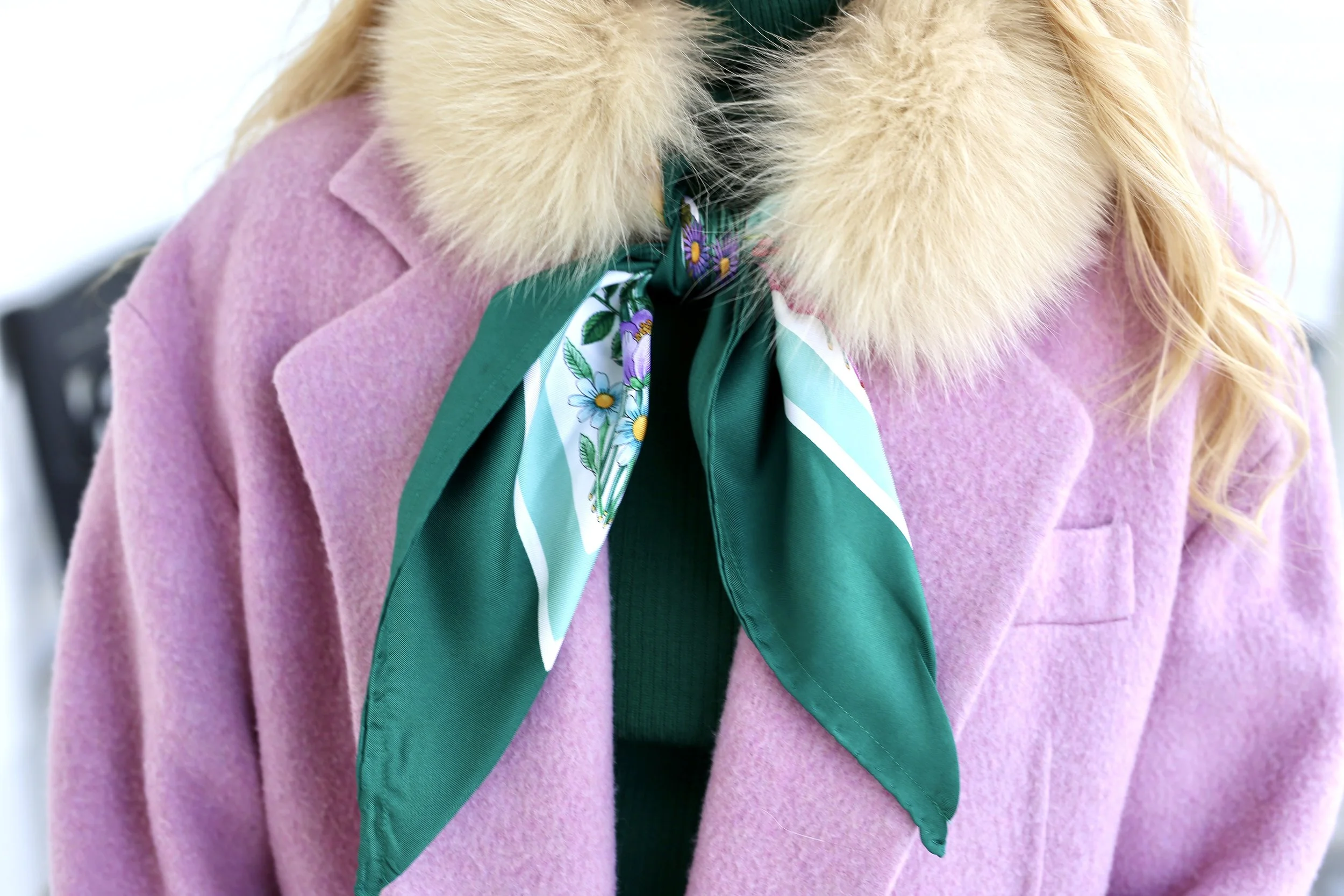 Close-up of a person wearing a pink coat with a faux fur collar and a green silk scarf with floral patterns around the neck.