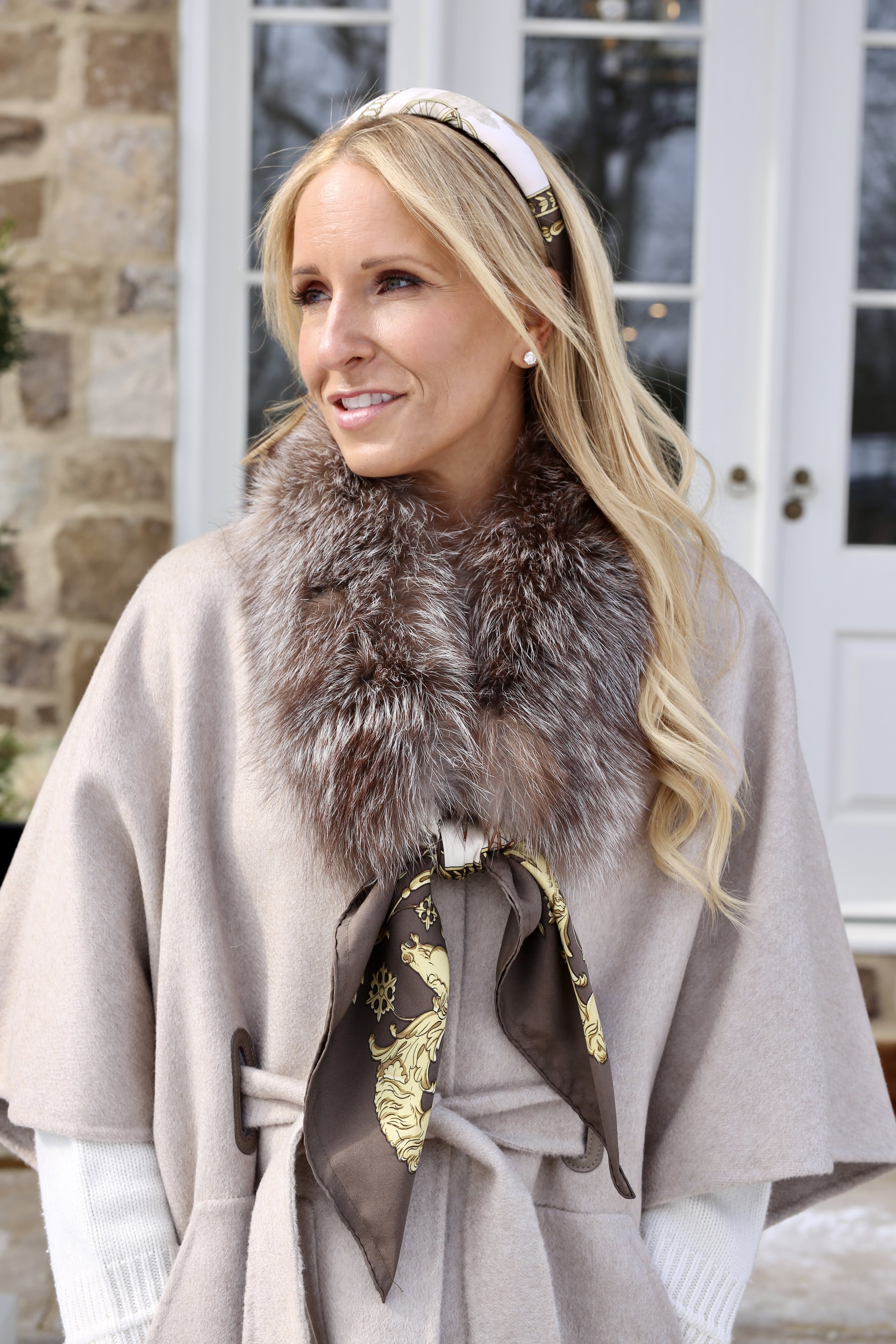 A woman wearing a beige coat with a fur collar and a decorative scarf tied around her neck, standing outside near a white door and stone wall.