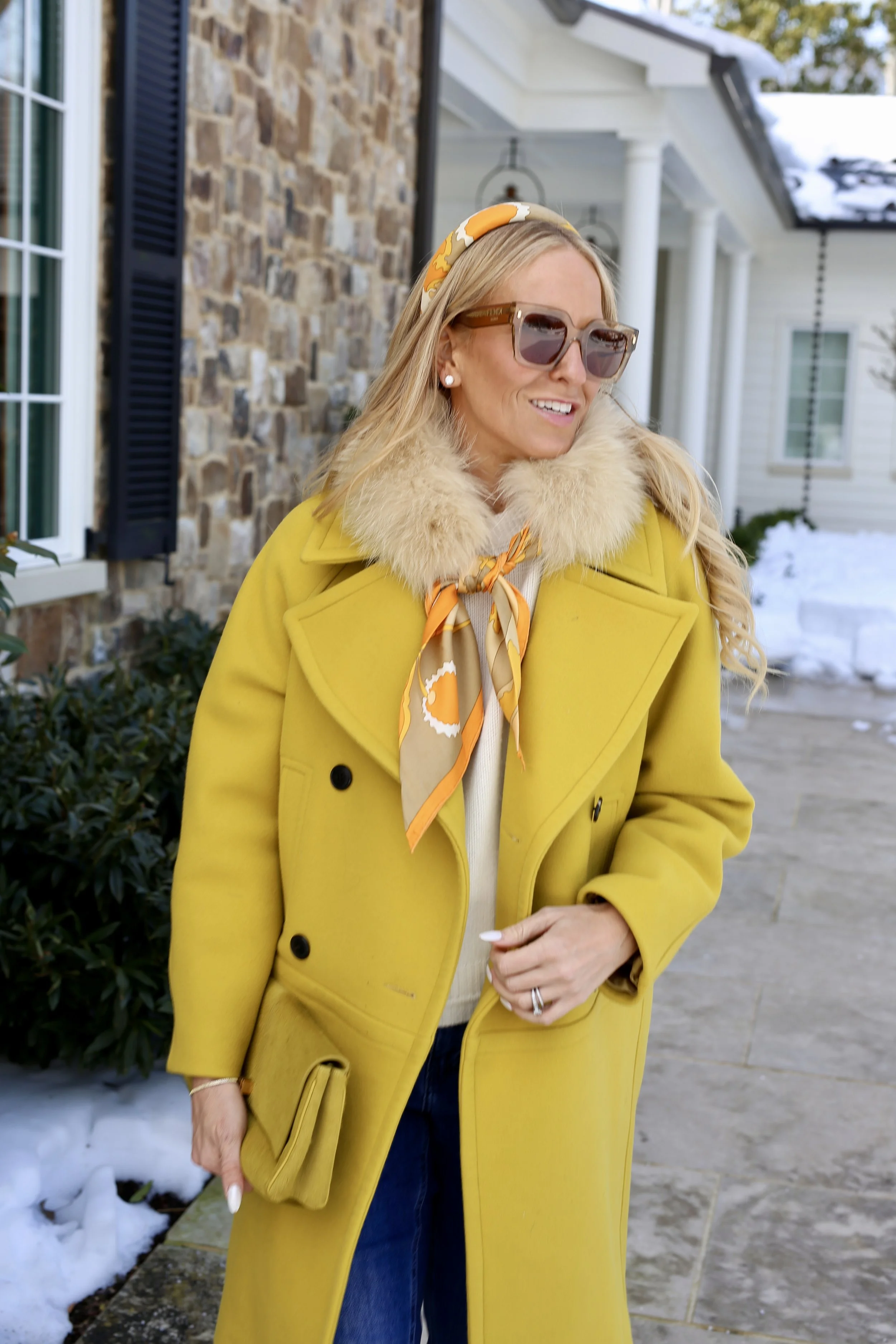 A woman with blonde hair wearing sunglasses, a yellow coat with a fur collar, and a colorful scarf tied around her neck, standing outdoors in winter near a house with stone and siding exterior.