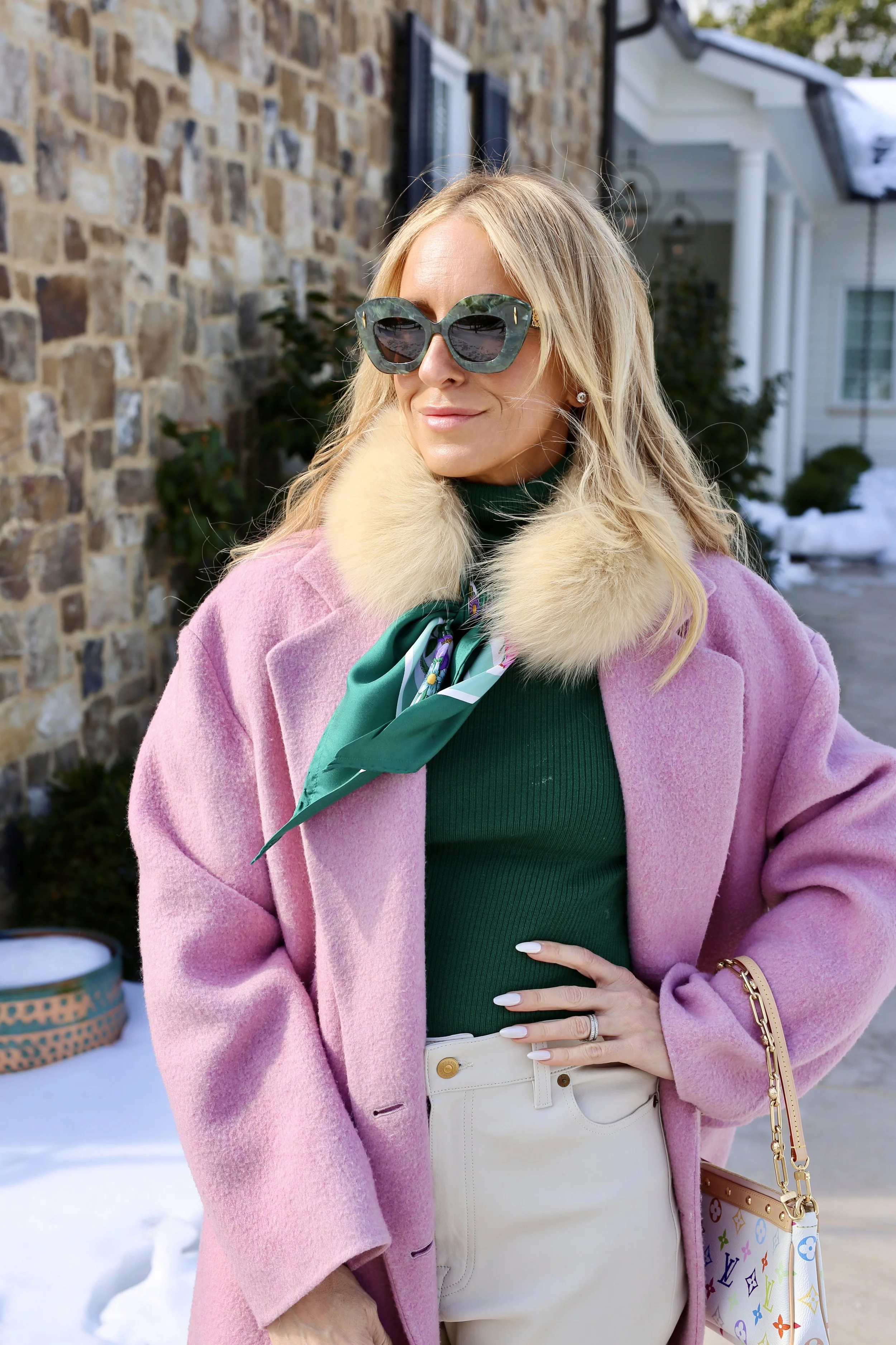 A woman with blonde hair wearing oversized sunglasses, a pink coat with a fur collar, a green turtleneck, beige pants, and a colorful Louis Vuitton handbag, standing outside in a snowy residential area.