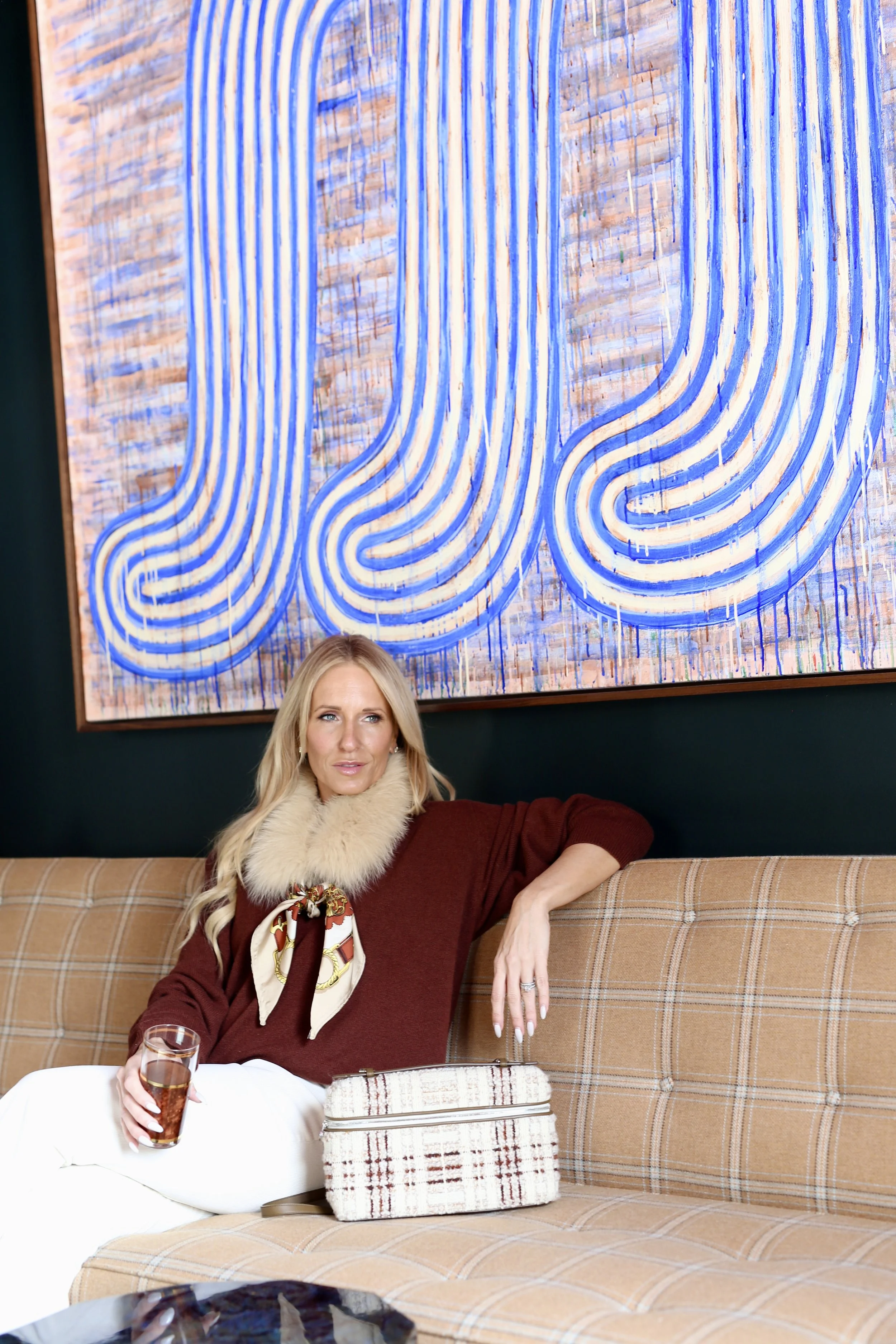 A woman with blonde hair wearing a brown sweater, a furry scarf, and a patterned silk scarf, sitting on a plaid beige couch holding a glass of drink, with a cream-colored purse placed beside her. Behind her is a large abstract artwork with blue and beige lines.