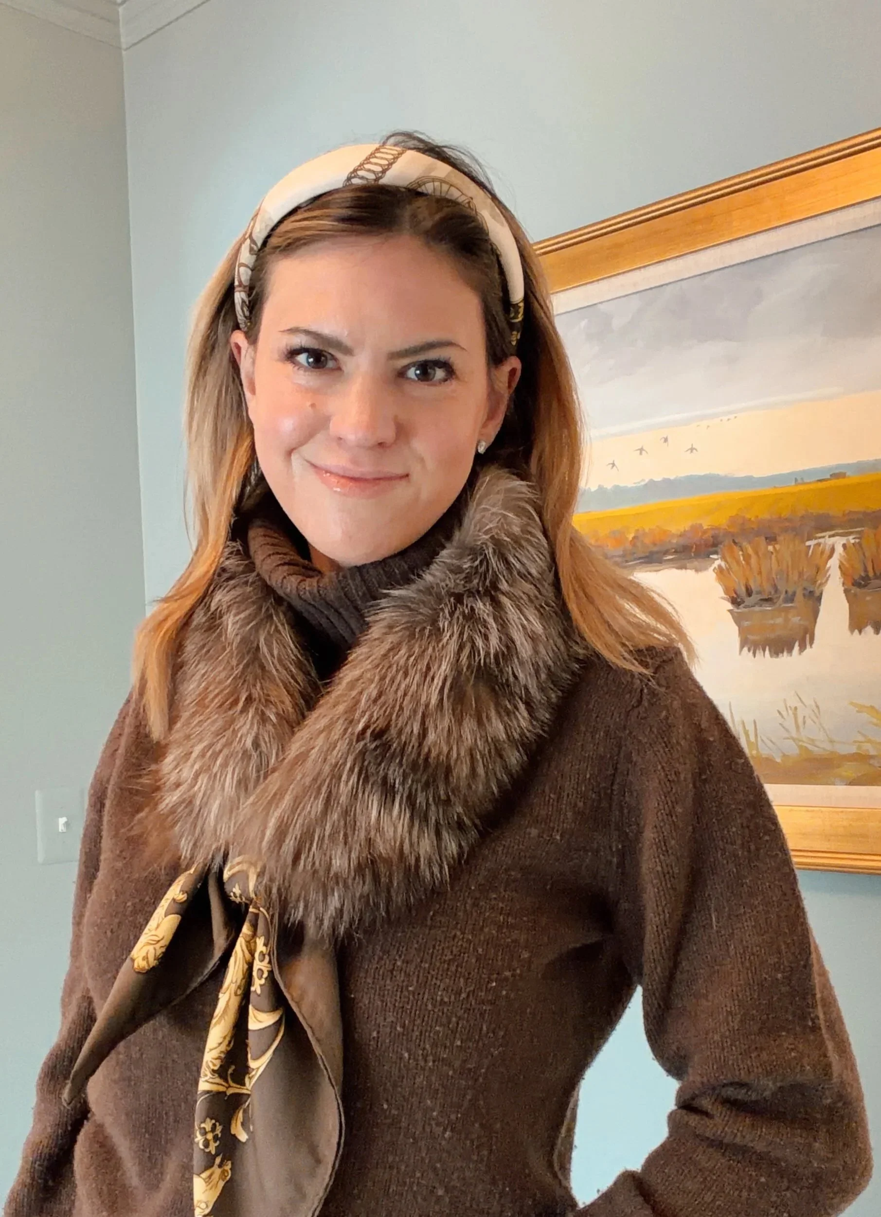 A woman with light skin and long, light brown hair, wearing a brown coat with a fur collar, a patterned scarf, and a headband, standing indoors in front of a landscape painting with a lake, trees, and a cloudy sky.
