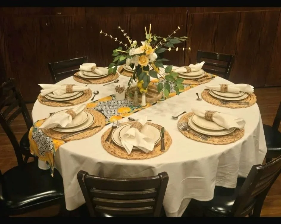 Round dining table set with white tablecloth, place settings with white plates, napkins, and silverware on woven placemats. In the center, there is a floral arrangement with yellow and white flowers and greenery.