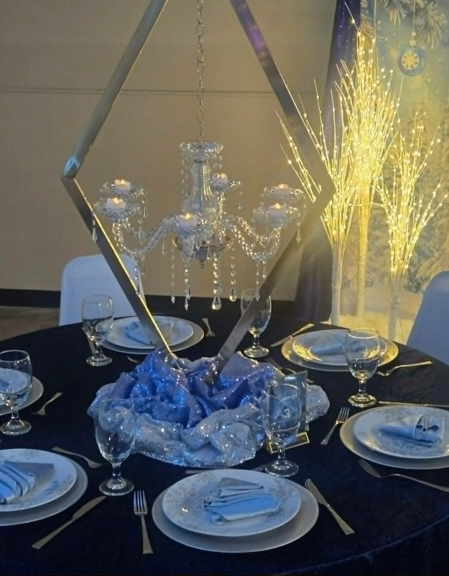 Elegant dining table set with white plates, silverware, wine glasses, white napkins, and a crystal chandelier hanging above. The table has a dark tablecloth and a decorative centerpiece with a metallic frame and fabric in shades of blue and silver. I