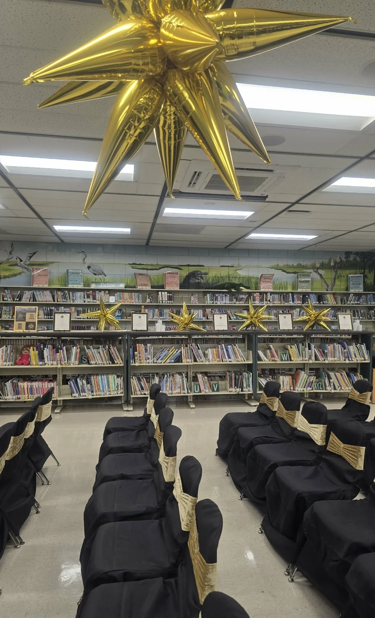 School library decorated with gold star-shaped balloons and chairs covered with black fabric and gold sashes, prepared for an event.