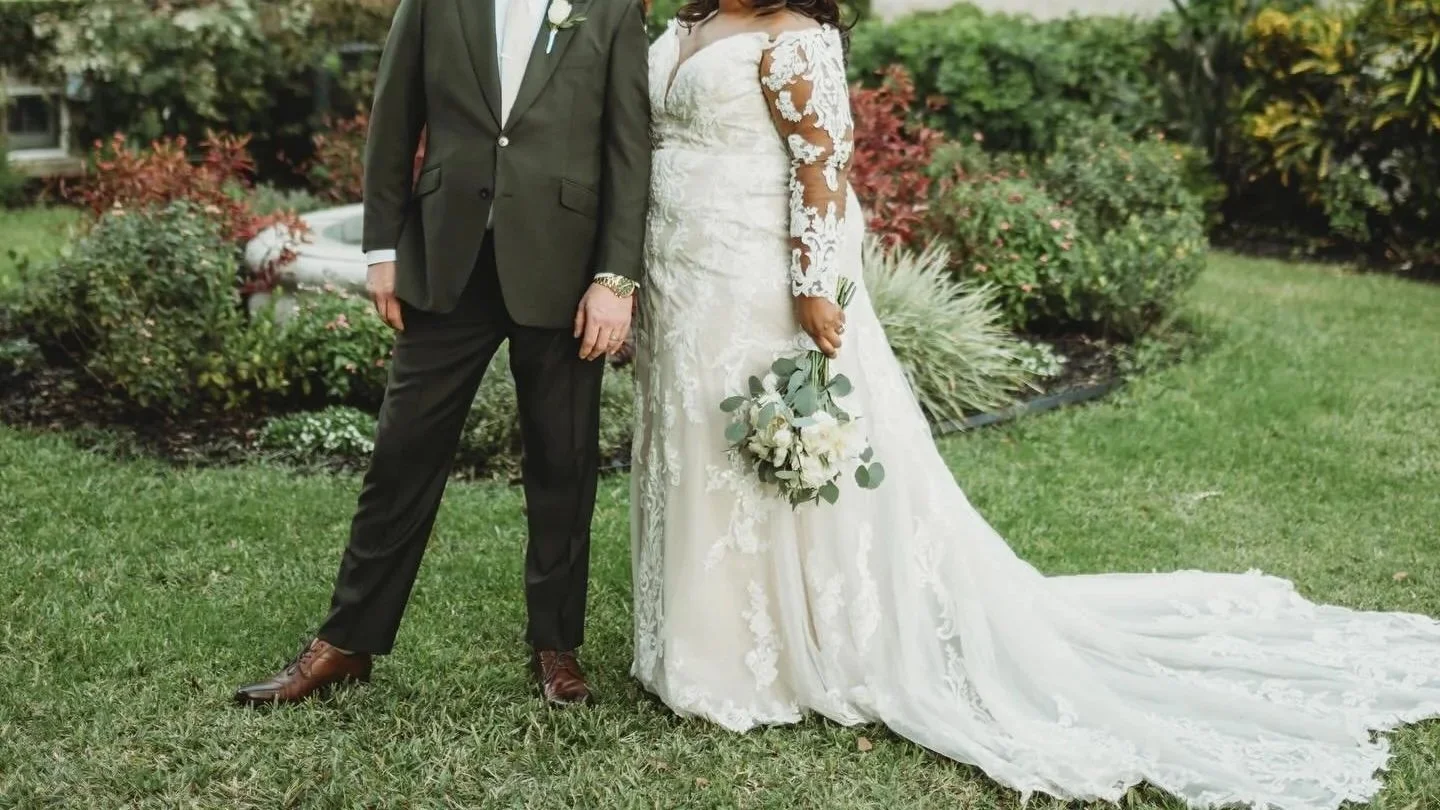A bride and groom standing outdoors on a grassy lawn. The groom is in a gray suit and brown shoes, while the bride is in a white lace wedding gown holding a bouquet of white and green flowers.