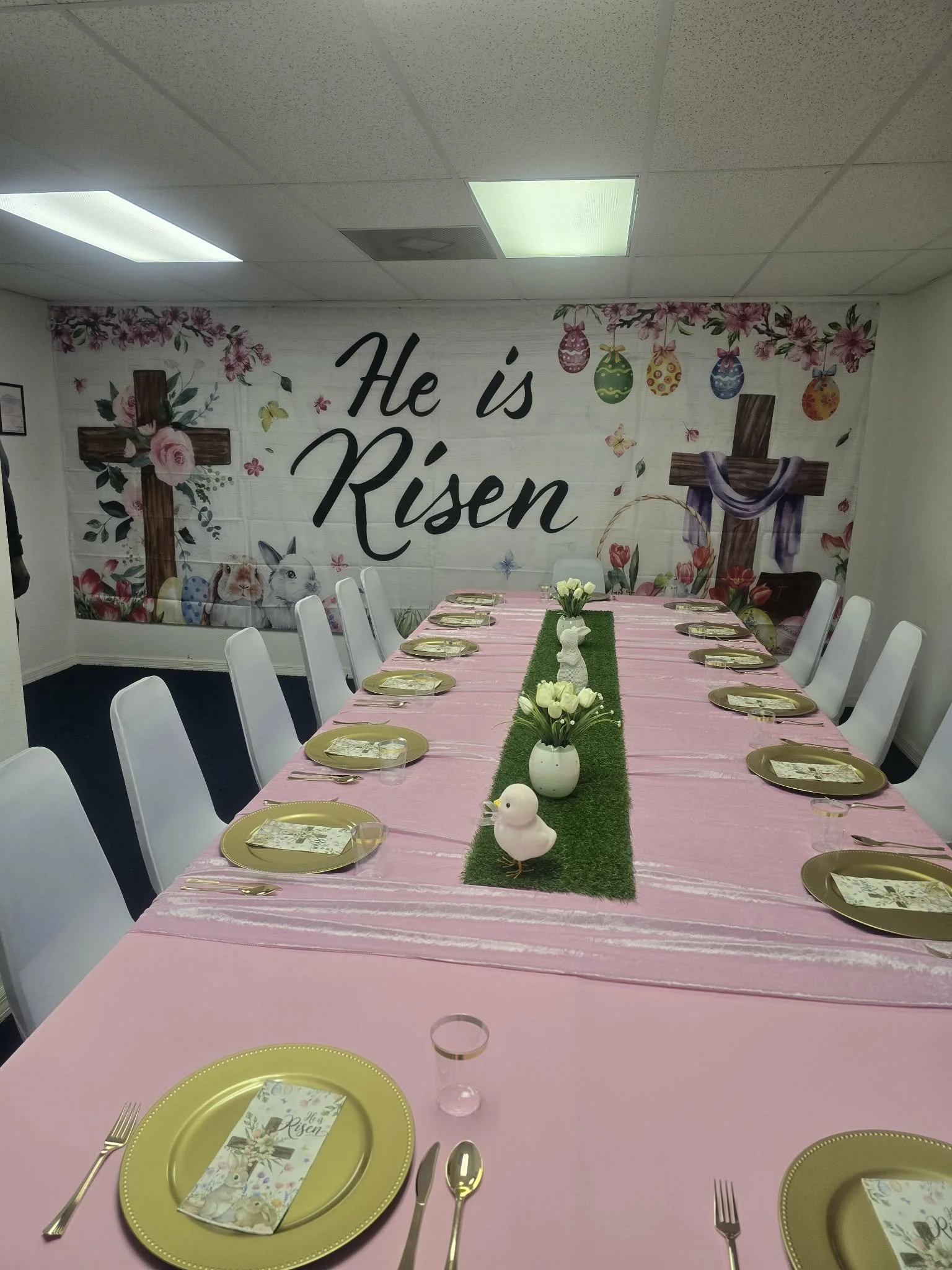 A long table decorated for Easter with pink tablecloth and gold plates, set with napkins, cups, and utensils, with a green grassy centerpiece featuring white flowers and a small white duck figurine in the middle. The background wall has a mural with 